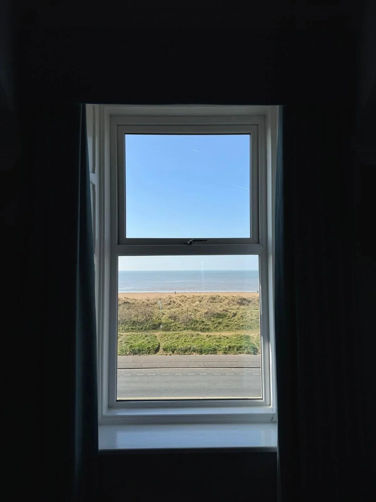 View through a window showing a sandy beach, grassy dunes, and the ocean under a blue sky.