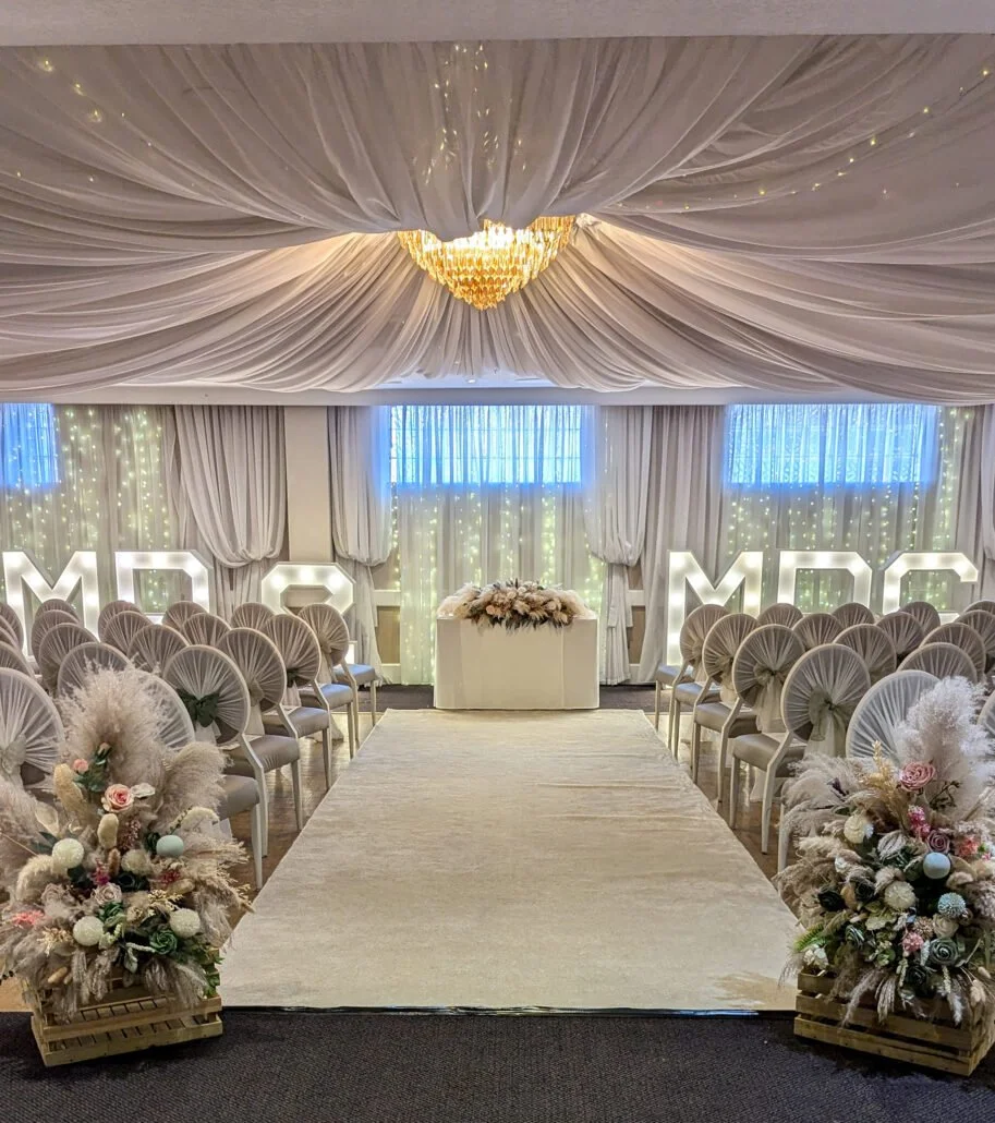 Elegant wedding ceremony setup with white draped ceiling, chandelier, and illuminated 'MDG' letters, floral arrangements, and rows of chairs.