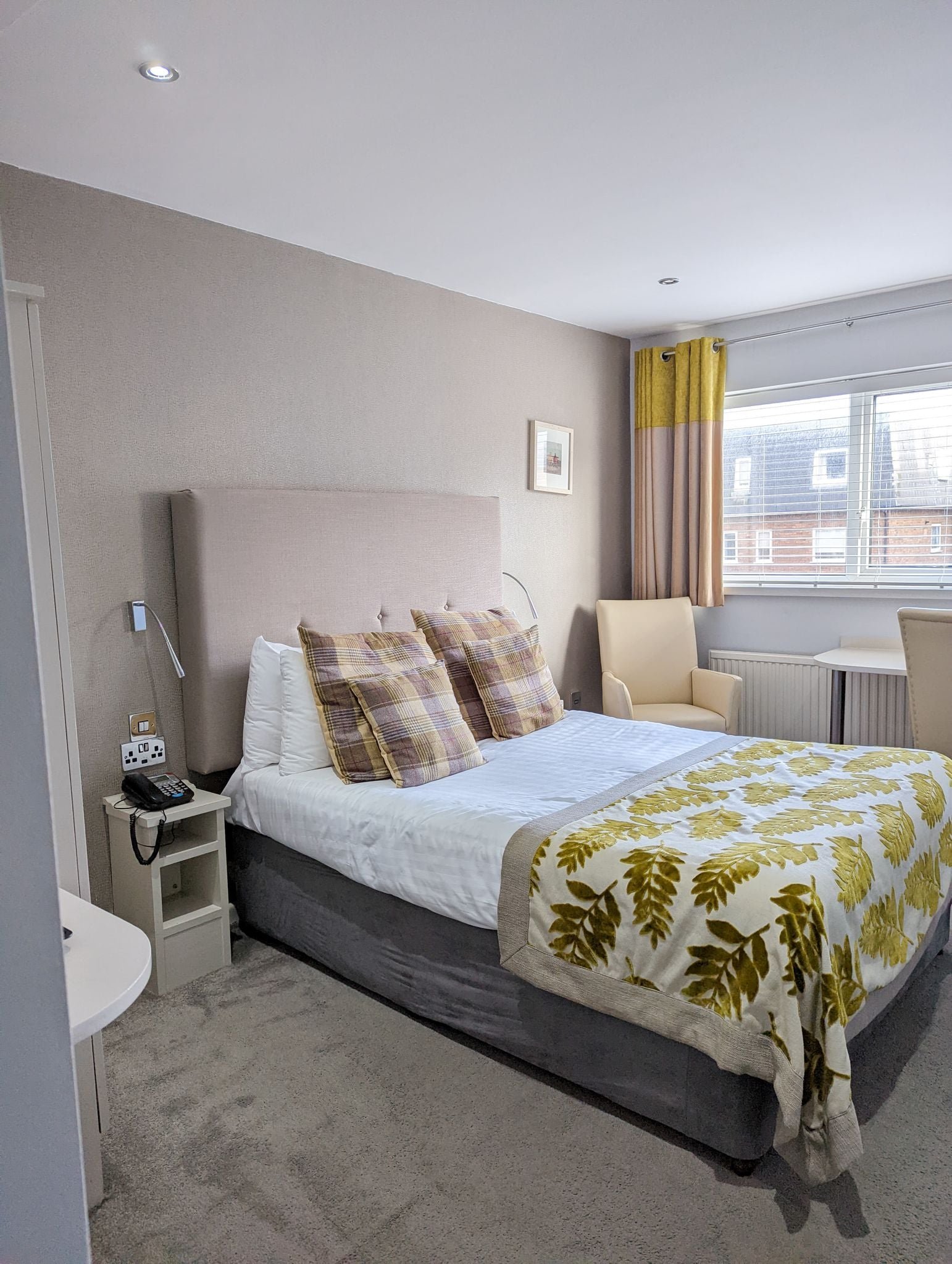 A neatly made hotel room bed with a beige upholstered headboard, plaid pillows, a white blanket with a yellow leaf pattern, and a gray bed base. To the left of the bed, there is a small nightstand with a telephone, a remote, and power outlets. The ro