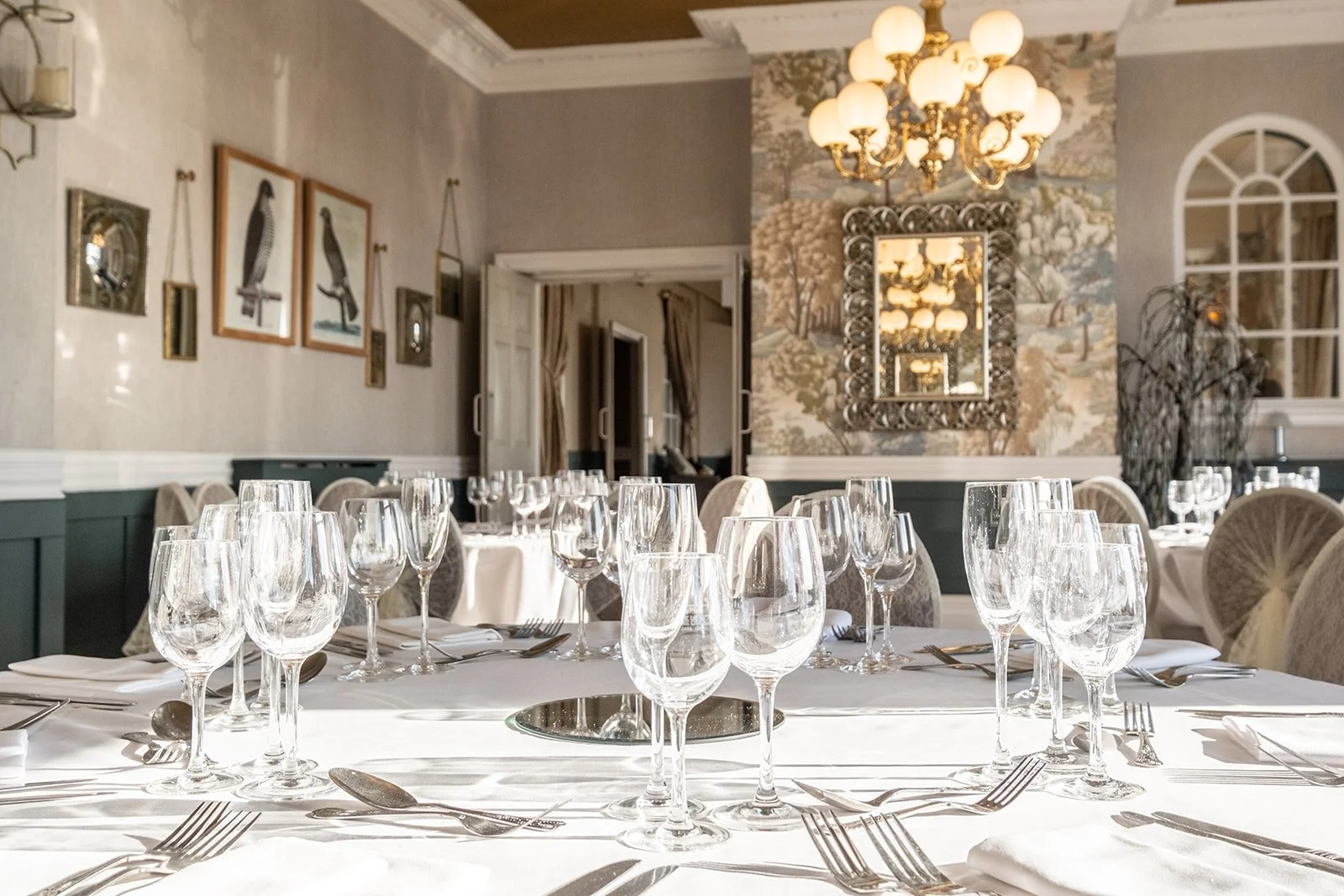 Elegant table setting in a well-decorated dining room with multiple glasses, silverware, white napkins, and a round mirror in the center of the table, with ornate wall art, a chandelier, and decorative wallpaper in the background.