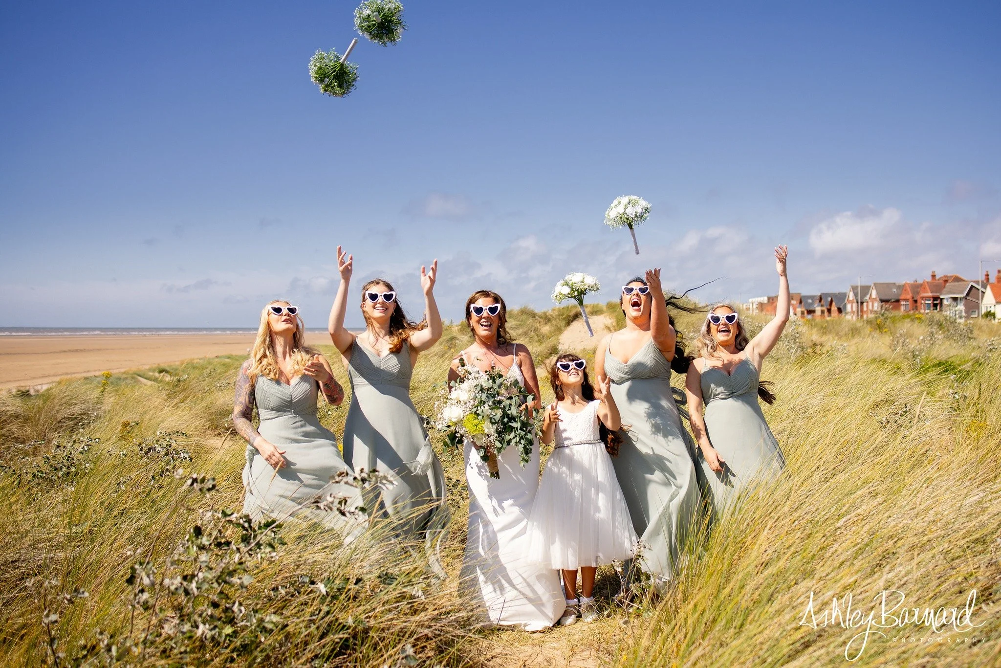 Bridesmaids and a young girl celebrating on a beach with sand dunes, throwing flower bouquets in the air on a sunny day.