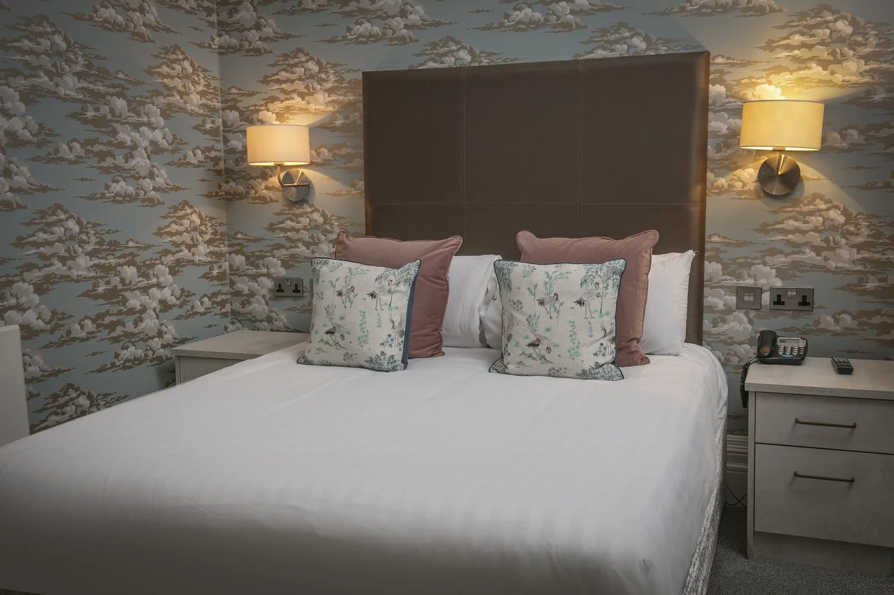 A hotel room with a bed, decorative pillows, and floral wallpaper, illuminated by wall-mounted lamps.