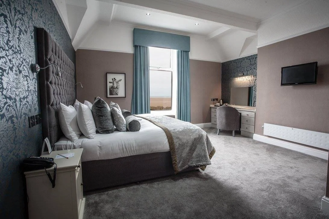 A hotel room with a large bed, decorative pillows, a nightstand with a phone, a framed artwork, a window with blue curtains, a vanity with a mirror, and a wall-mounted TV.