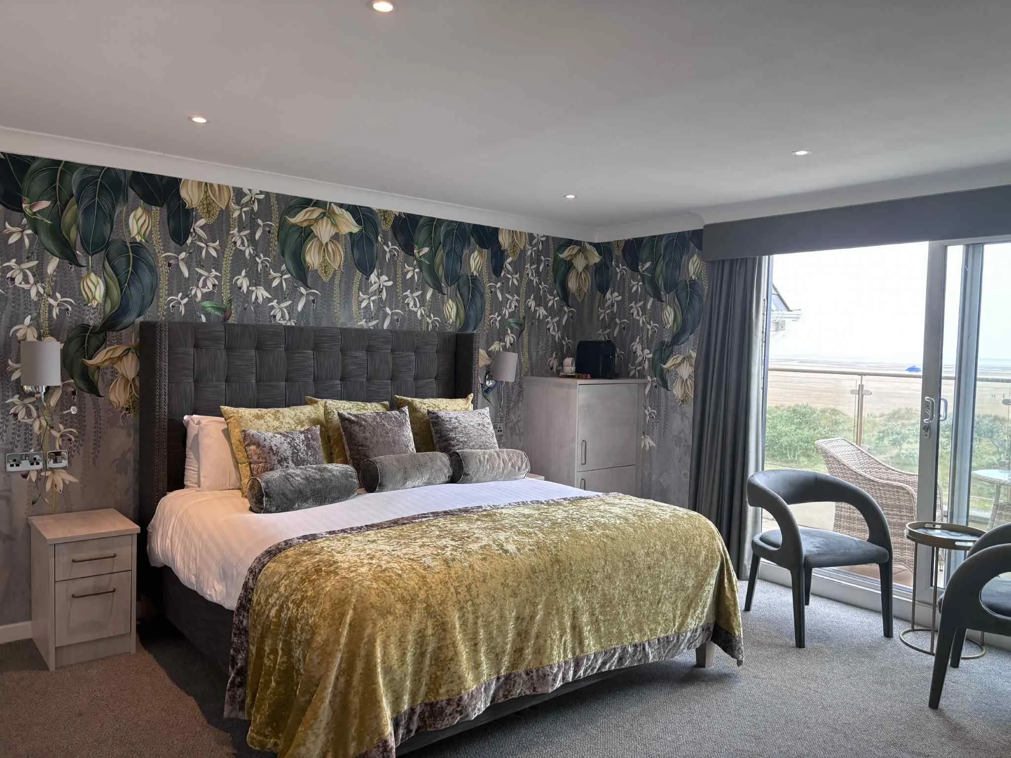 A modern hotel room with a large bed, decorative pillows, and a textured velvet bedspread. The room features a floral accent wall, bedside lamps, a window with a balcony, and outdoor seating.