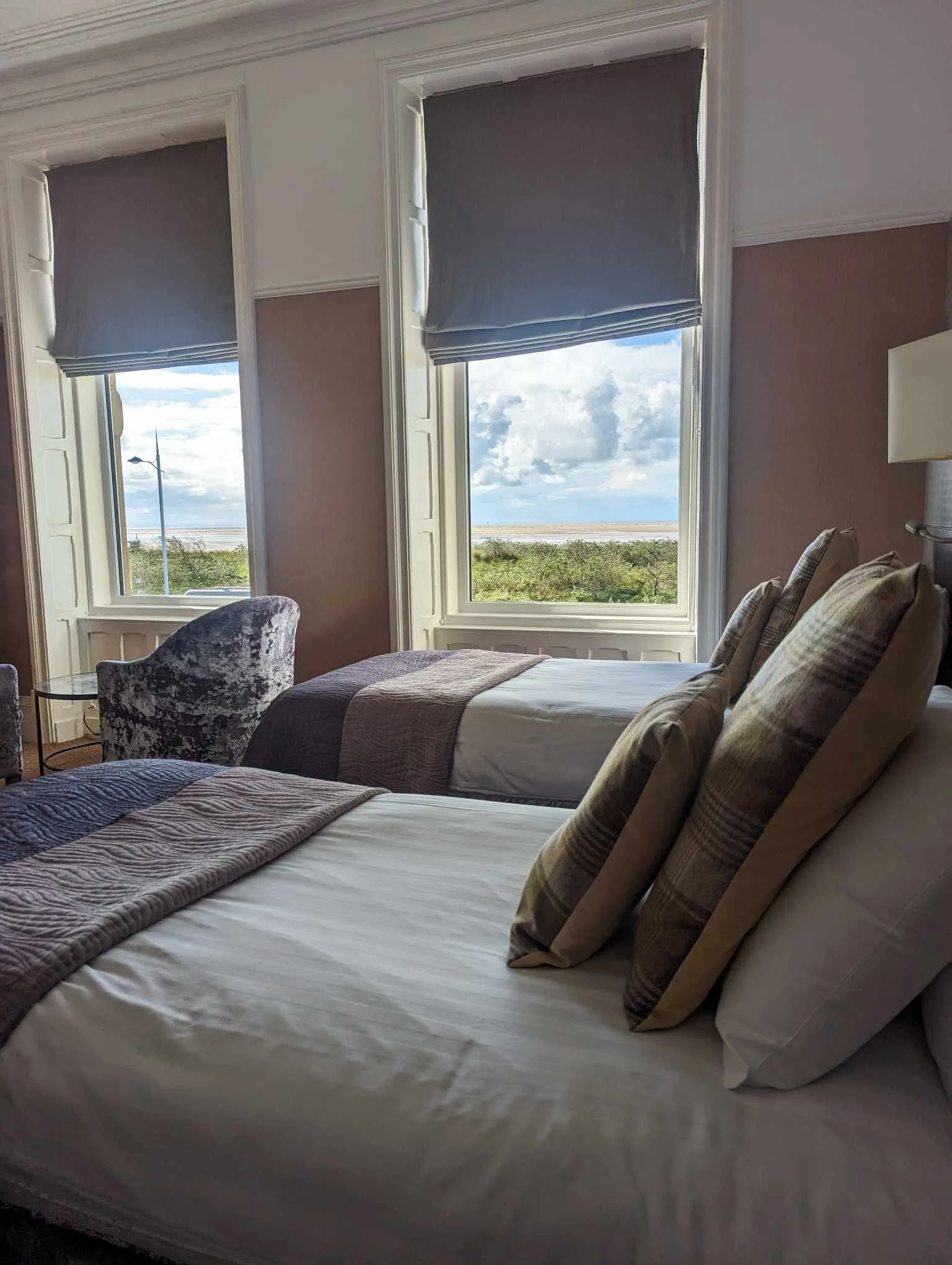 Two beds in a room with large windows showing a grassy landscape and cloudy sky outside, with blue window shades and patterned pillows.