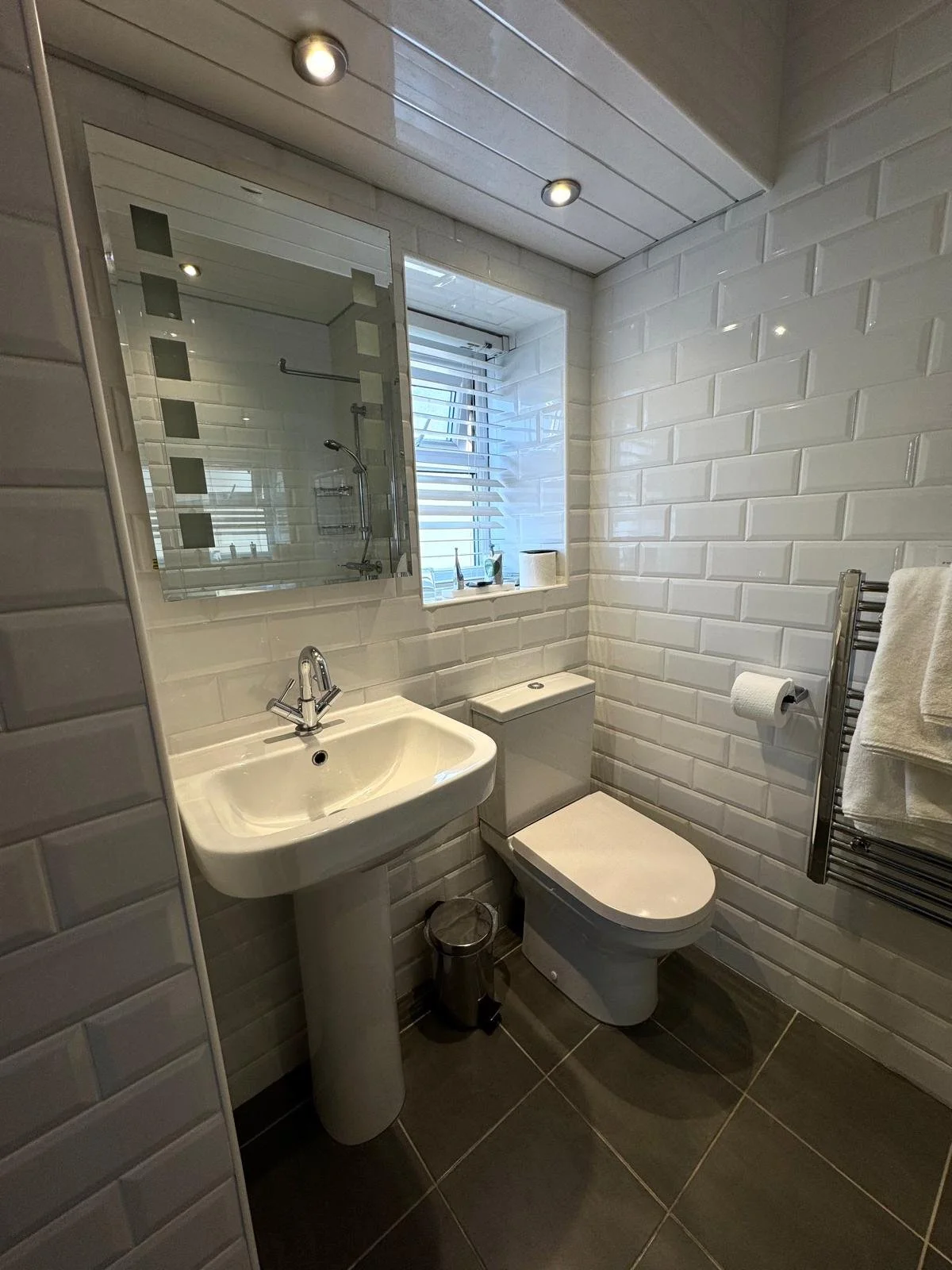 A small bathroom with white tile walls, a window with blinds, a pedestal sink, a toilet, a mirror above the sink, a stainless steel towel rack with towels, a small trash bin, and a shower visible through a glass partition in the reflection.