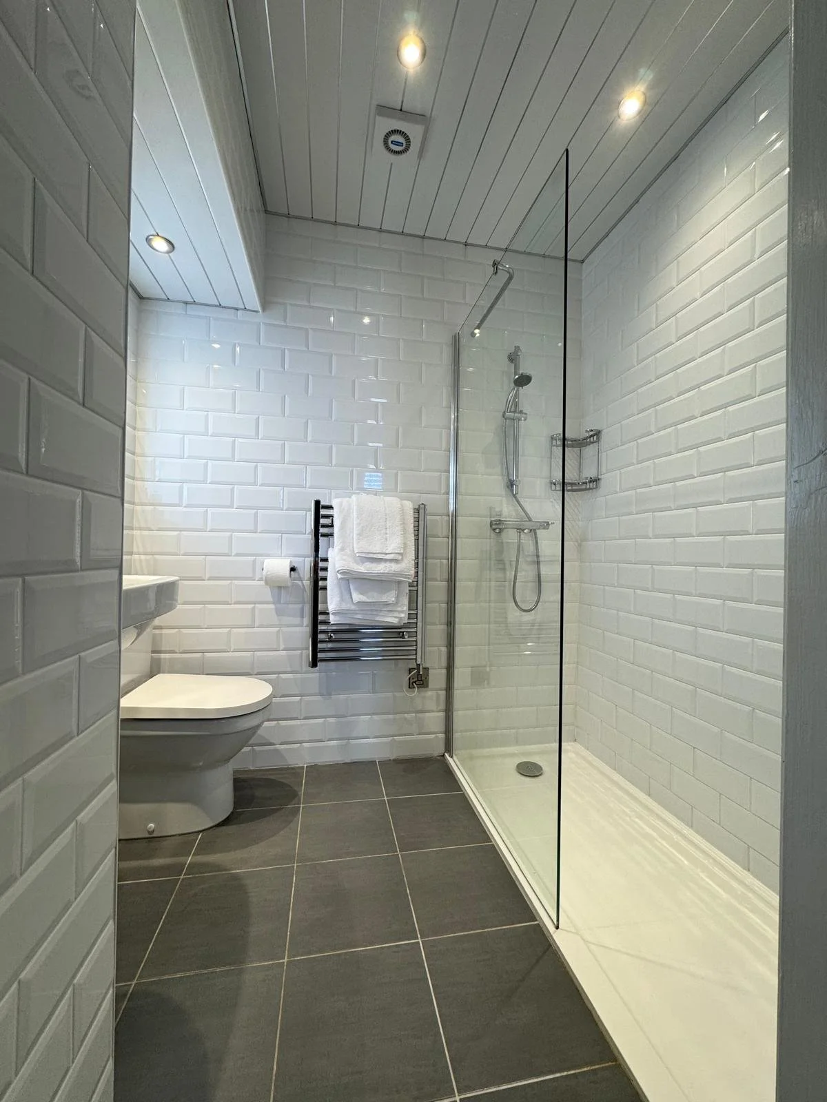Modern bathroom with white subway tile walls, dark floor tiles, a walk-in shower with clear glass door, a towel rack with white towels, a toilet, and a white ceiling with recessed lighting.