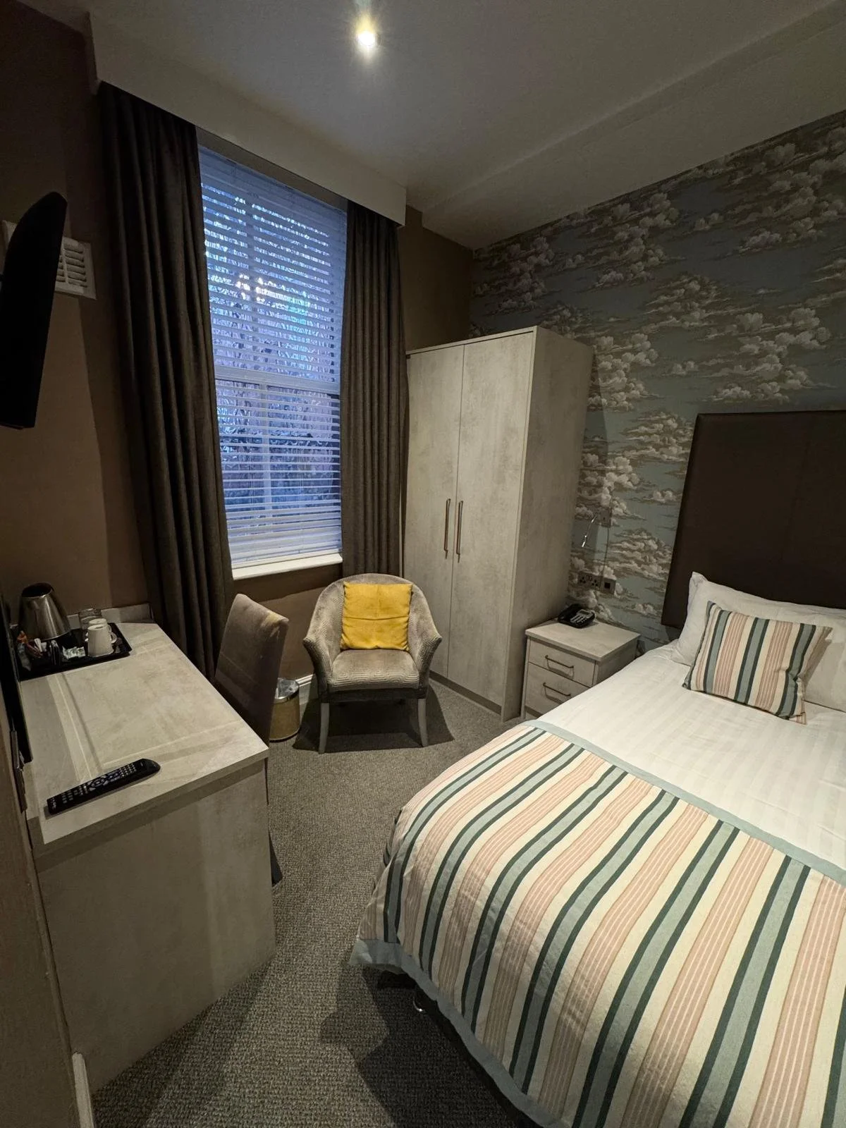 A hotel room with a bed, a window with dark curtains, a small chair with a yellow pillow, a wardrobe, a nightstand with a telephone, and a dresser with a TV, all decorated with patterned wallpaper.