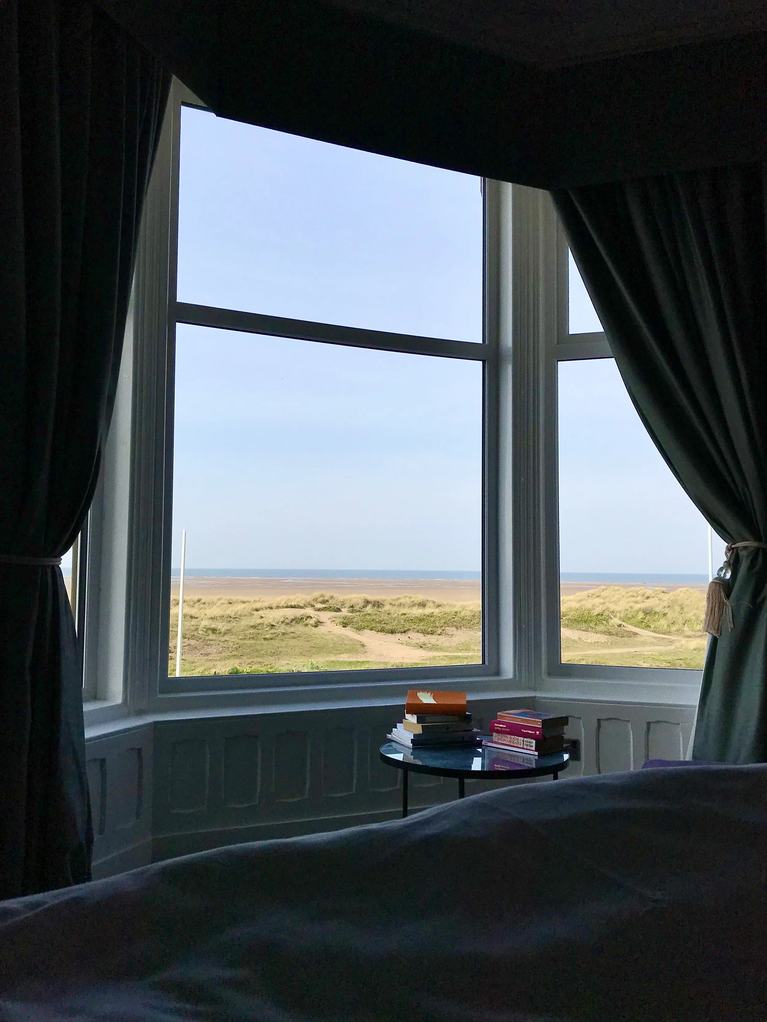 View of coastal dunes through a large bay-window with dark curtains, and a small table with stacks of books inside a room.