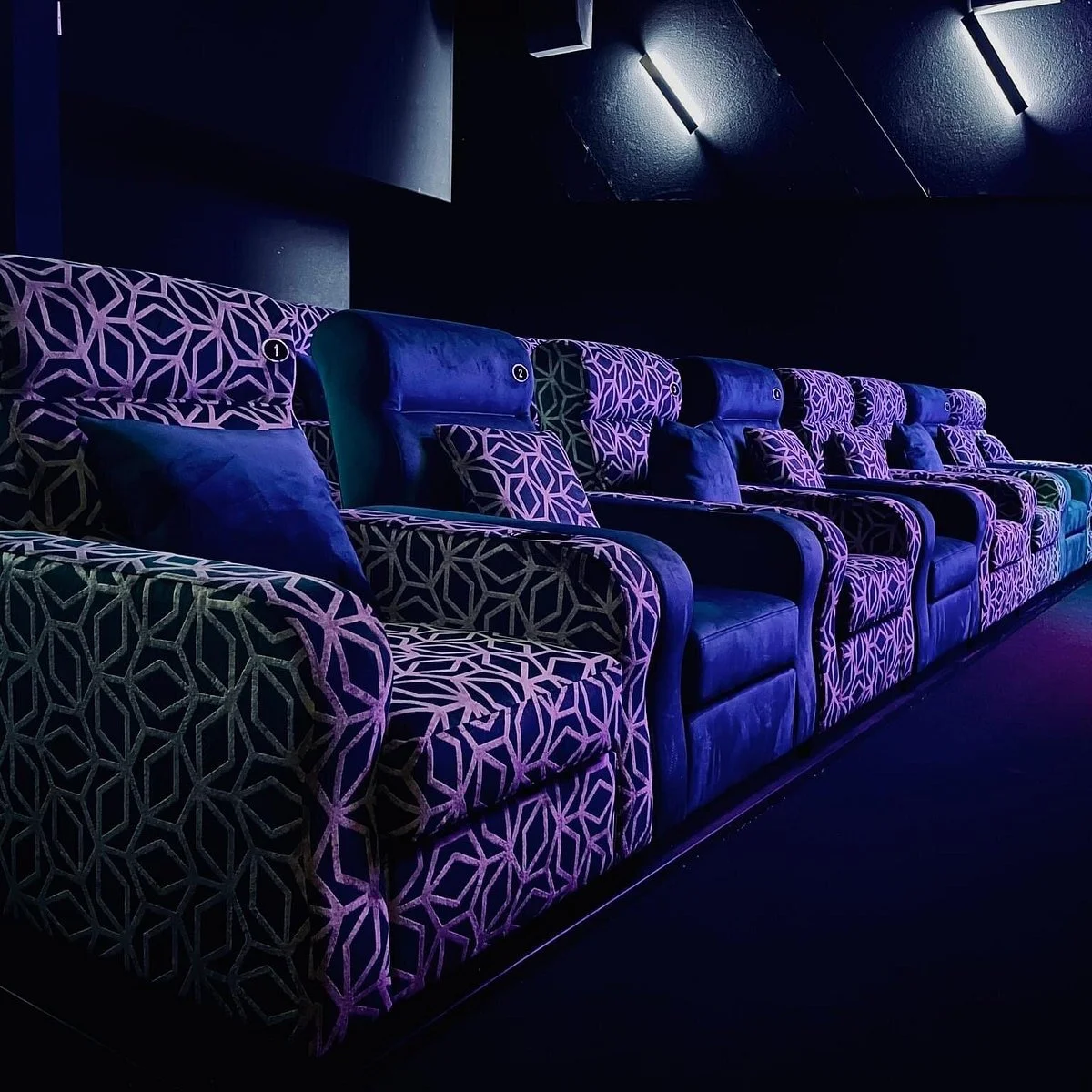 A row of plush, patterned theater seats with purple and black geometric designs and matching pillows, set in a dimly lit room with dark walls and soft lighting.