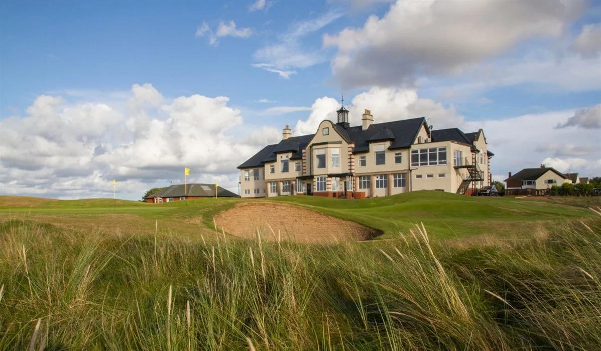 Large house on a golf course with green grass, sand trap, and flagpoles under a partly cloudy sky.