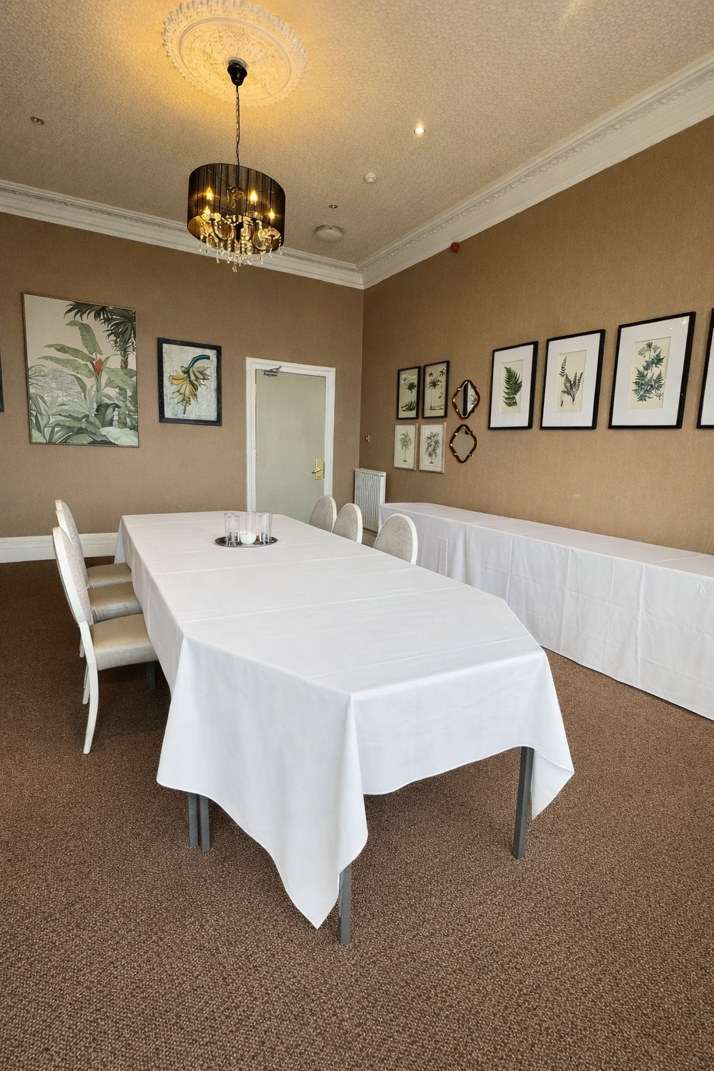 A dining room with a long table covered with a white tablecloth, seven cream-colored chairs, wall art of botanical prints, a chandelier, and a side table or buffet against the back wall.
