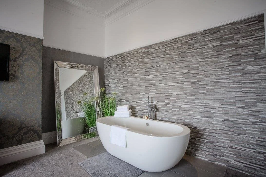 Modern bathroom with a white freestanding bathtub, a large mirror leaning against the wall, and decorative plants. The room features a textured gray tile wall and a plush gray rug.