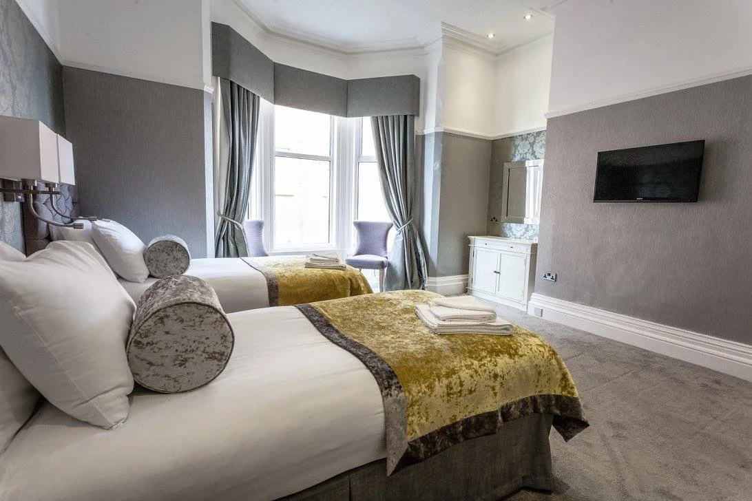Two neatly made twin beds with white linens, cylindrical pillows, and gold and gray blankets in a hotel room with gray walls, a large bay window with curtains, a wall-mounted TV, and a white cabinet.