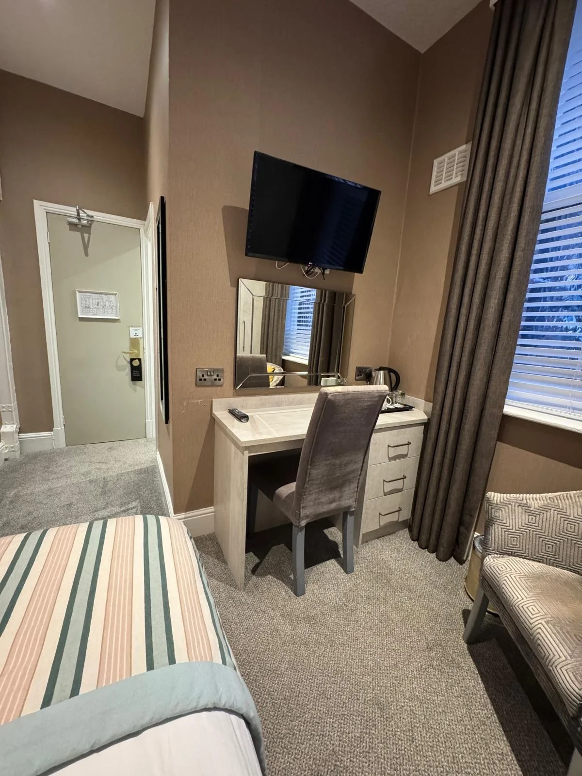 View of a hotel room corner with a bed, a desk with a chair, a mirror, a wall-mounted TV, a window with closed blinds, and curtains.