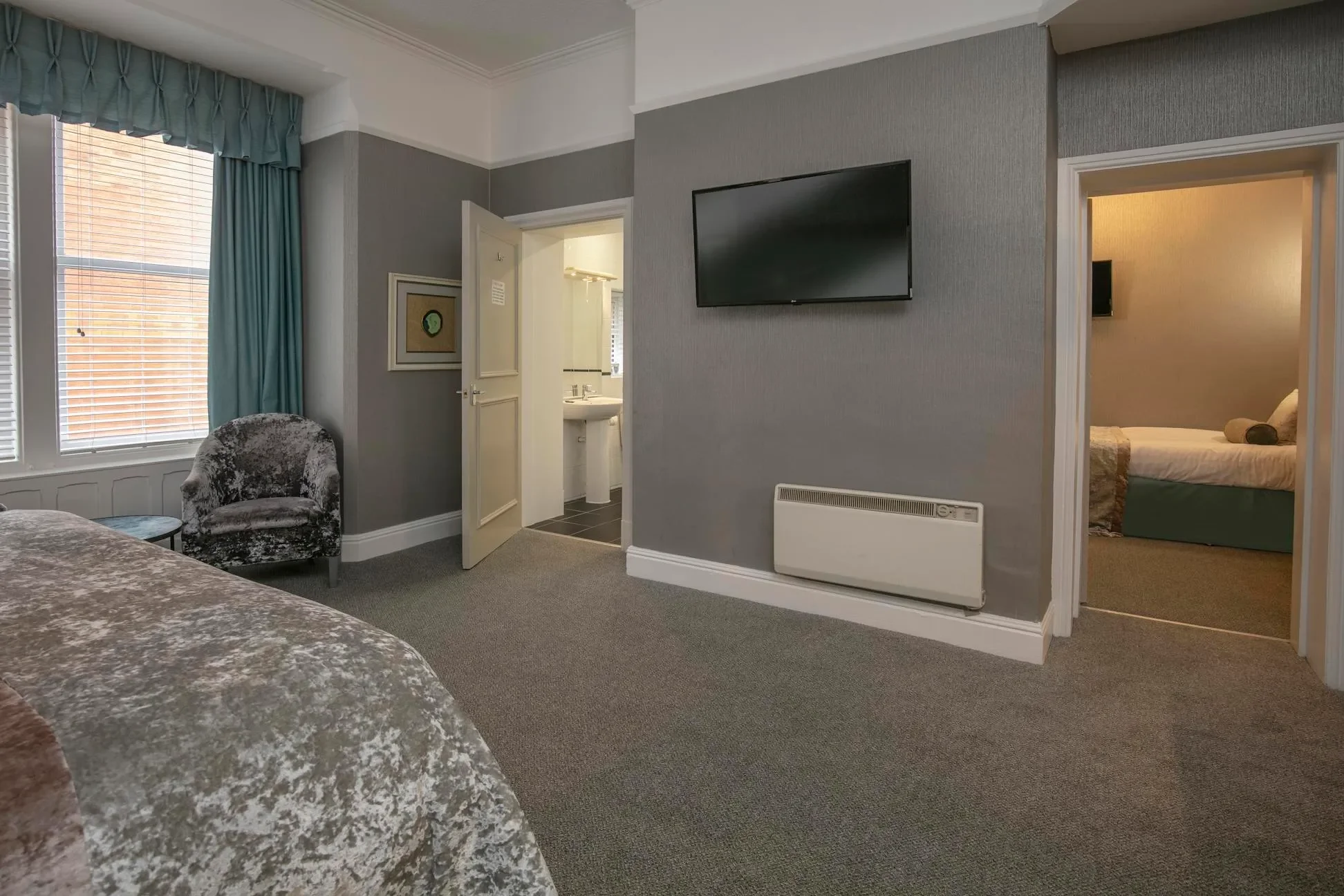 Hotel room with gray walls, patterned bedspread, a velvet armchair near windows with blue curtains, flat-screen TV on wall, open door to bathroom, and doorway to a second bedroom with twin beds.