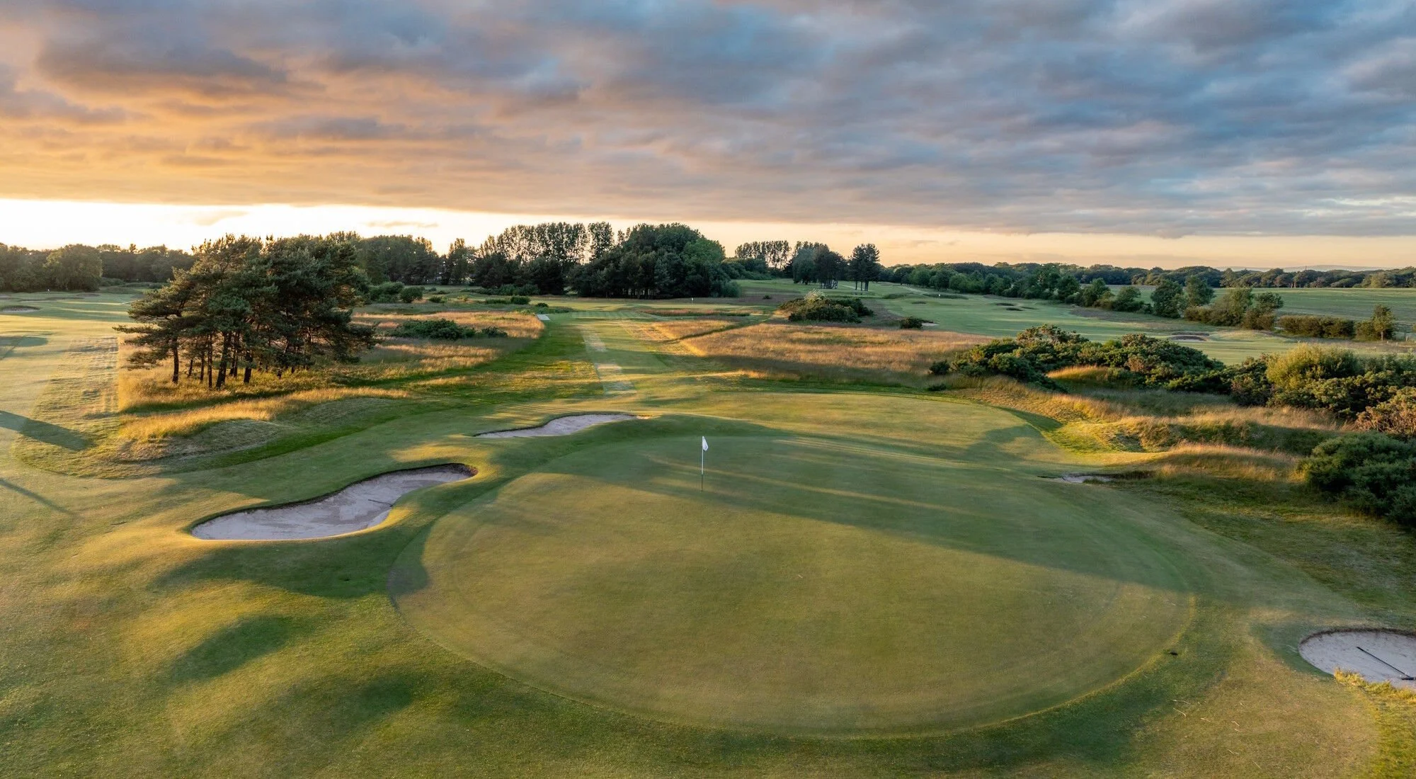 An expansive golf course at sunset with green fairways, sand bunkers, and a flag on the putting green in the foreground, surrounded by trees and rolling hills under a partly cloudy sky.
