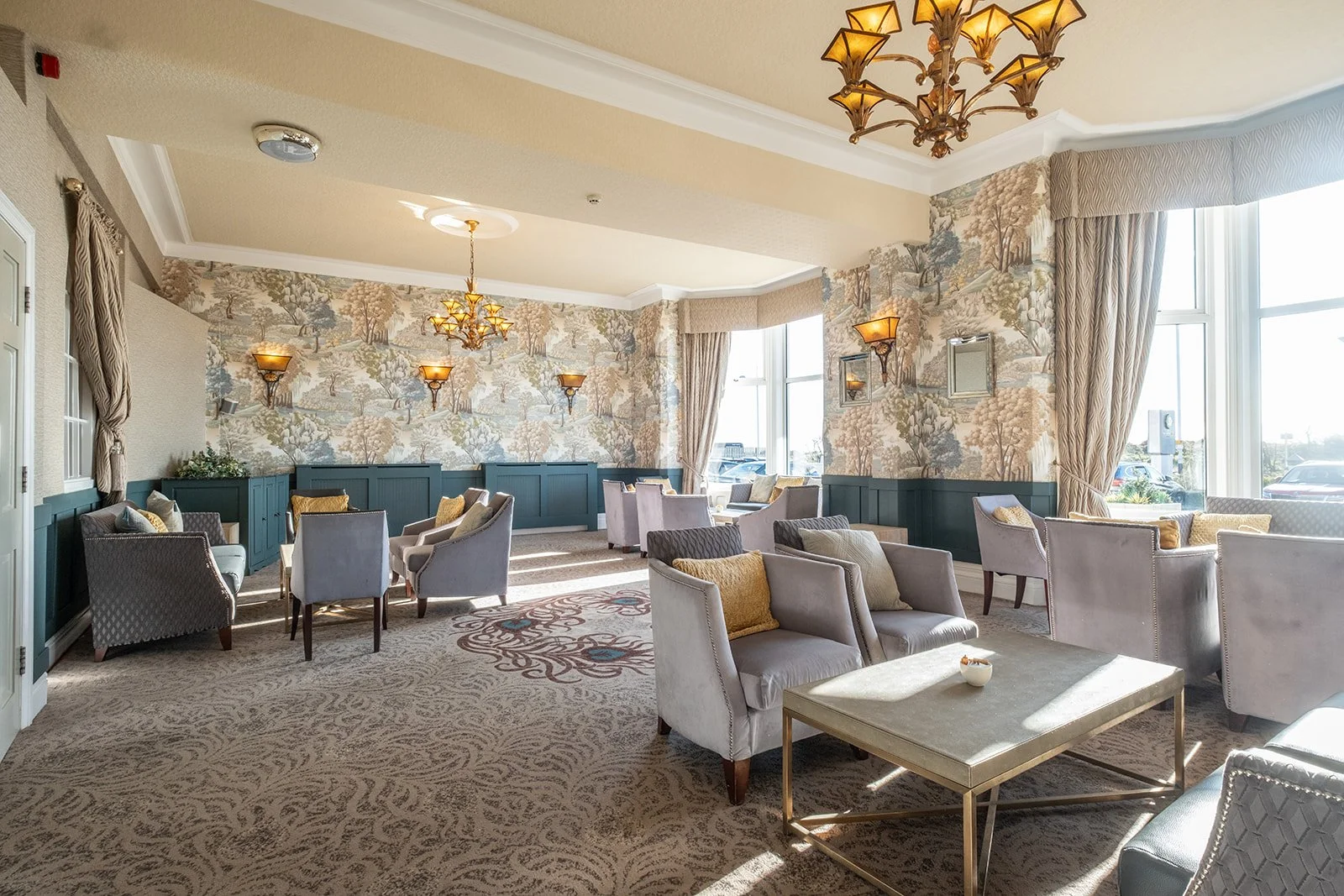 Sunlit hotel lounge with beige patterned carpet, pastel armchairs, and floral wallpaper. Chandeliers and wall sconces provide lighting, with large windows letting in natural sunlight.