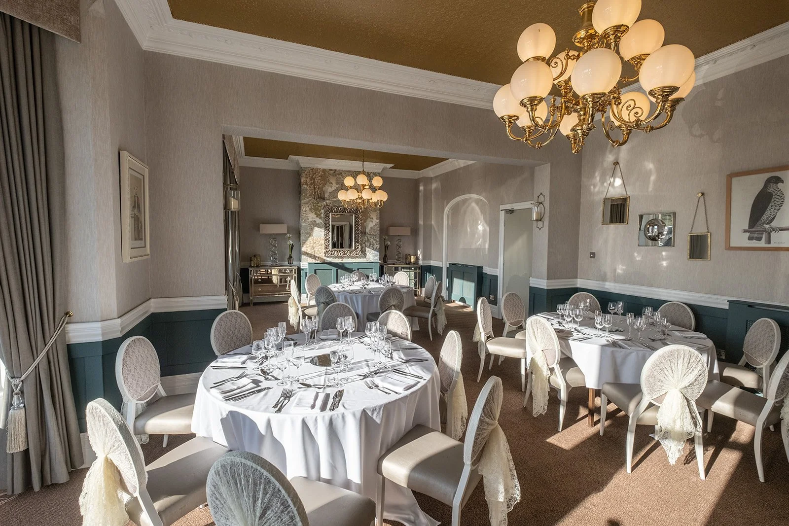 Elegant dining room with round tables covered in white tablecloths, set with glassware and silverware, surrounded by chairs with lace covers, in a room with beige walls, a gold chandelier, and wall art.