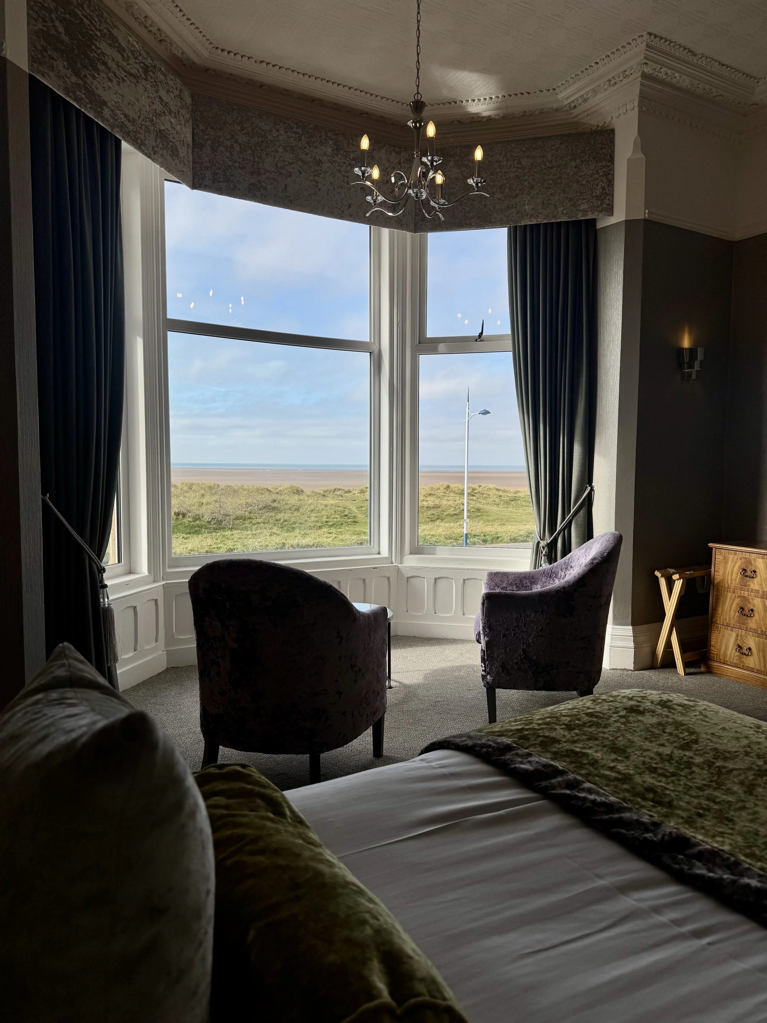 Hotel room with a large bay window, two purple velvet chairs, a bed with a green velvet bedspread, and a view of a grassy area and the ocean outside.