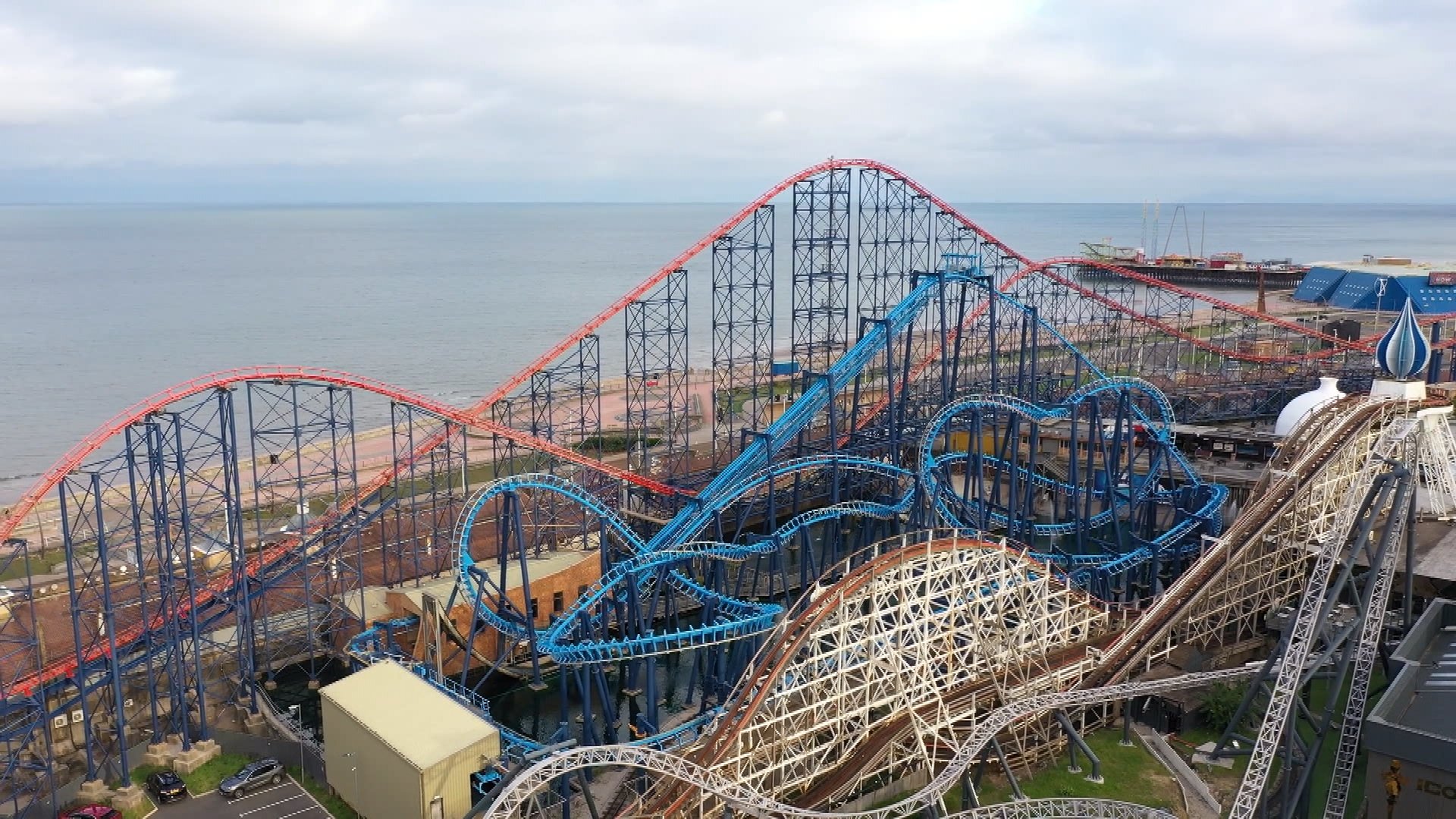 An amusement park with multiple roller coasters near the ocean, featuring various vibrant tracks, some blue and brown, with a cloudy sky overhead.