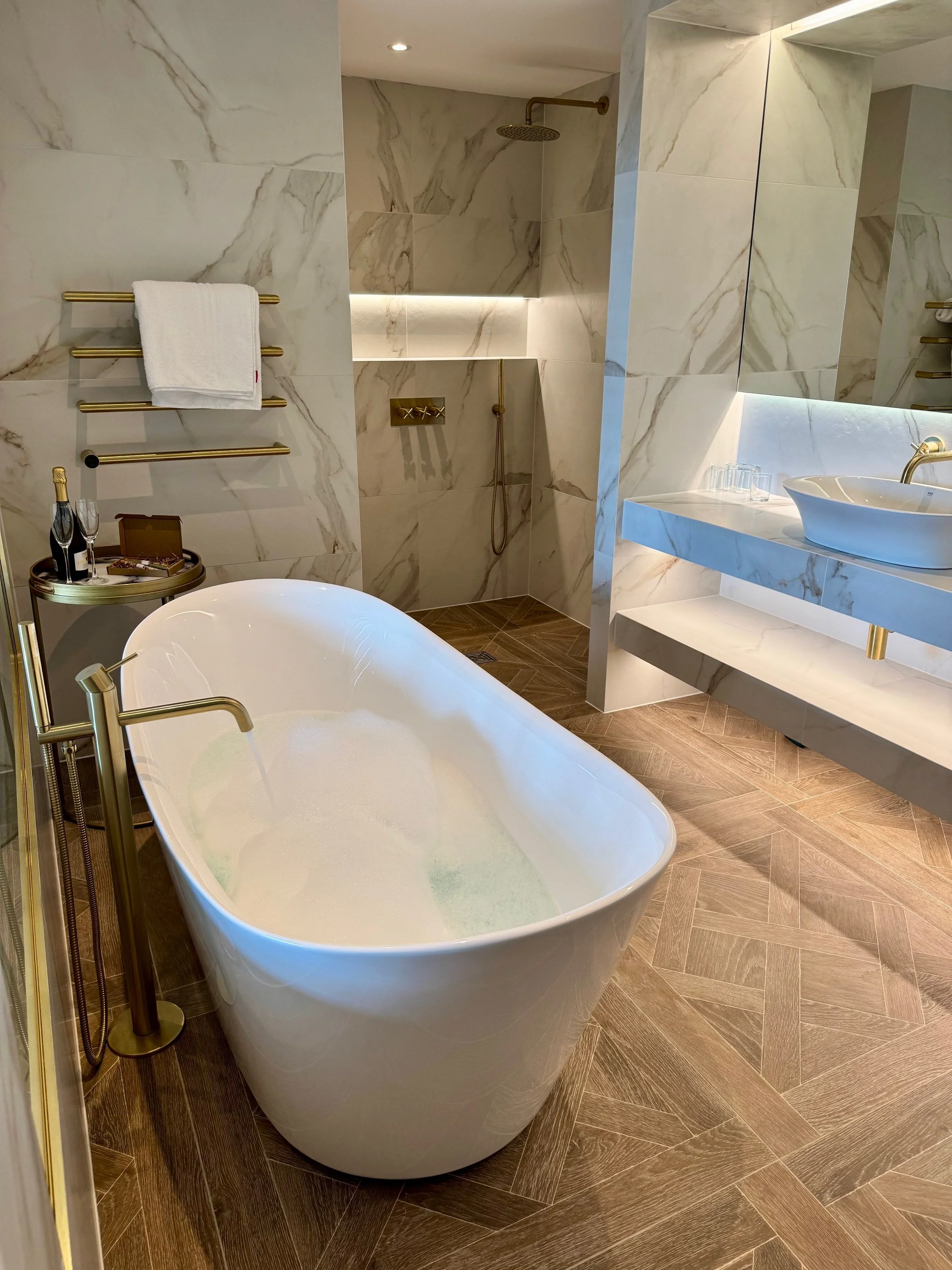 Luxury bathroom with a freestanding bathtub filled with bubbles, a walk-in shower with a rainfall showerhead, marble walls, a white sink on a marble countertop, and wooden flooring.