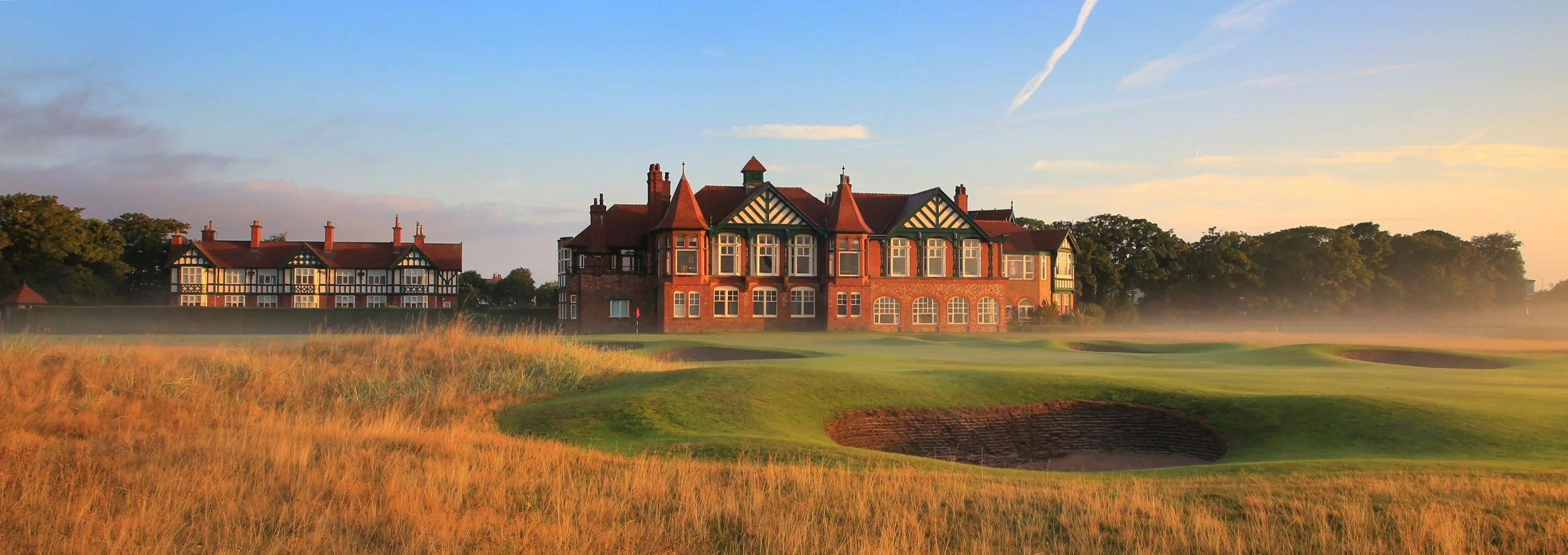 A large, historic brick mansion with multiple towers and gabled roofs, situated behind a golf course with sand bunkers and a putting green, during early morning or late afternoon with a sunrise or sunset in the background.