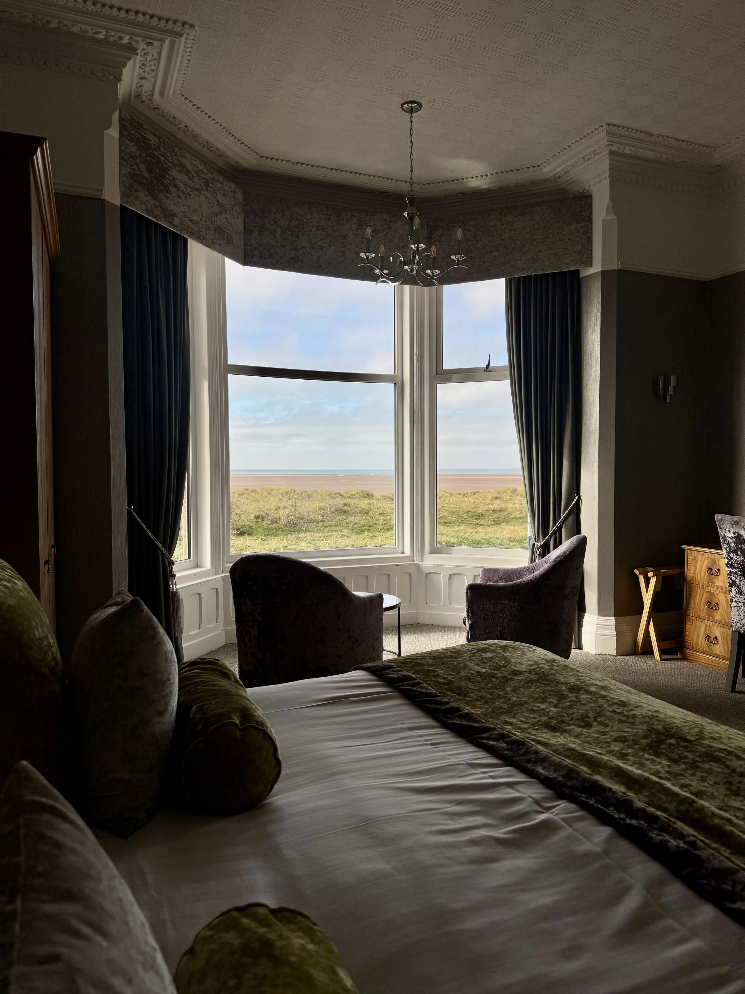 A bedroom with a large window overlooking a grassy landscape and cloudy sky. The room has dark curtains, a chandelier, a bed with green velvet pillows and a bedspread, two purple velvet armchairs near the window, and a wooden dresser.