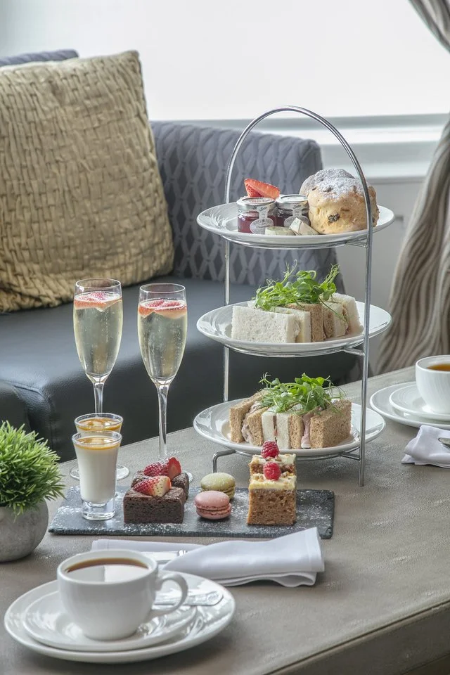 A three-tiered serving tray with assorted finger sandwiches, scones, and pastries, accompanied by glasses of champagne, a cup of coffee, and small desserts like macarons and cakes, set on a table with a white napkin and a small potted plant.