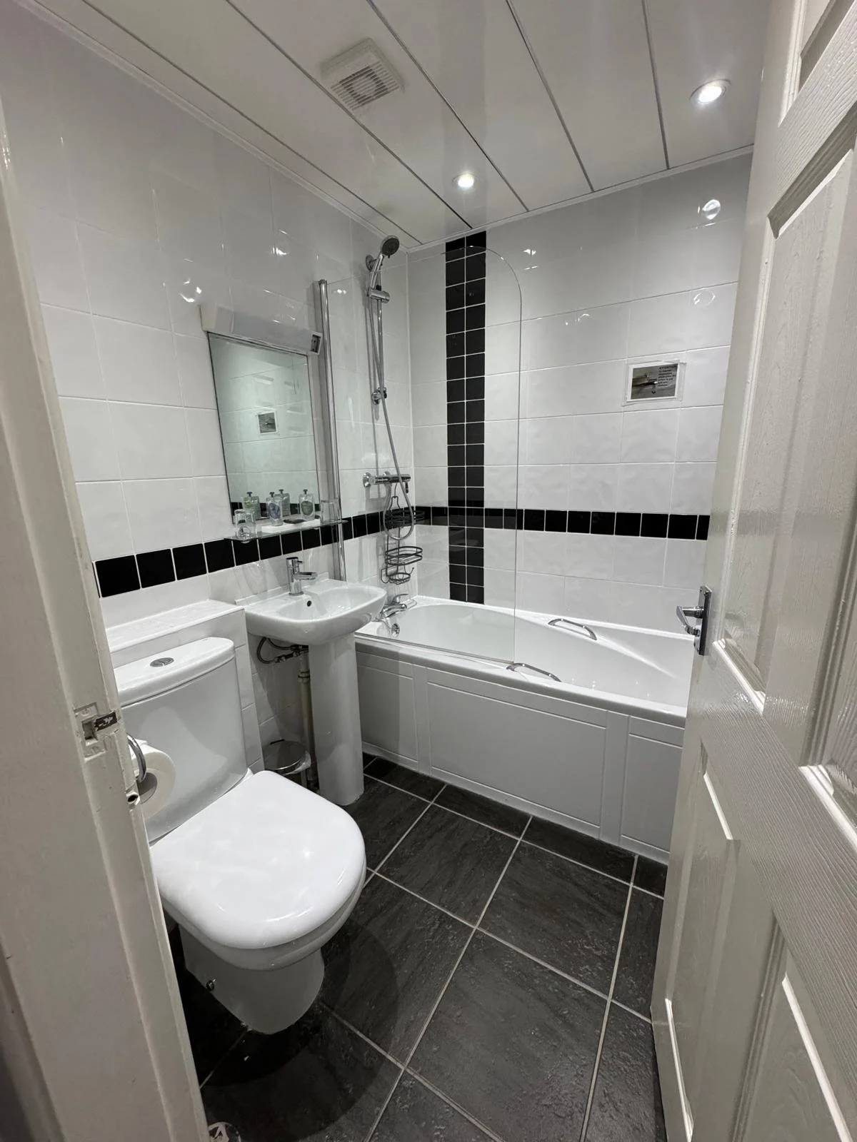 Small modern bathroom with white tiled walls, black accents, a toilet, a sink, a bathtub with a glass panel, and a showerhead.