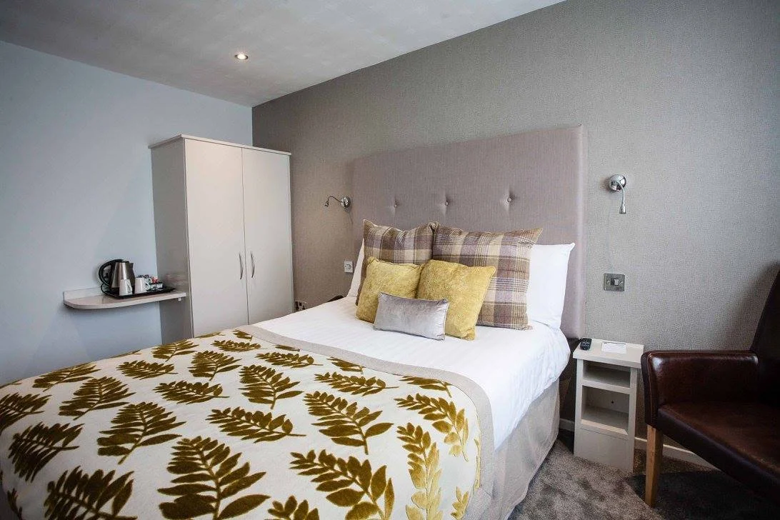 A neatly made bed with a beige upholstered headboard, multiple pillows, and a white and gold leaf-patterned blanket. To the right of the bed is a white nightstand with a remote and remote control. On the left side, a small shelf with a kettle and cups, and a white wardrobe are visible. The room has a gray and light blue wall, a gray carpet, and a brown armchair on the right.