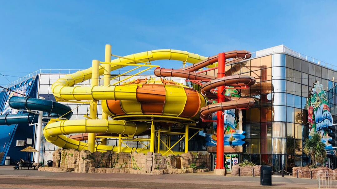 Water park with multiple water slides in yellow, brown, and red, outside a modern glass building with palm trees and a clear blue sky