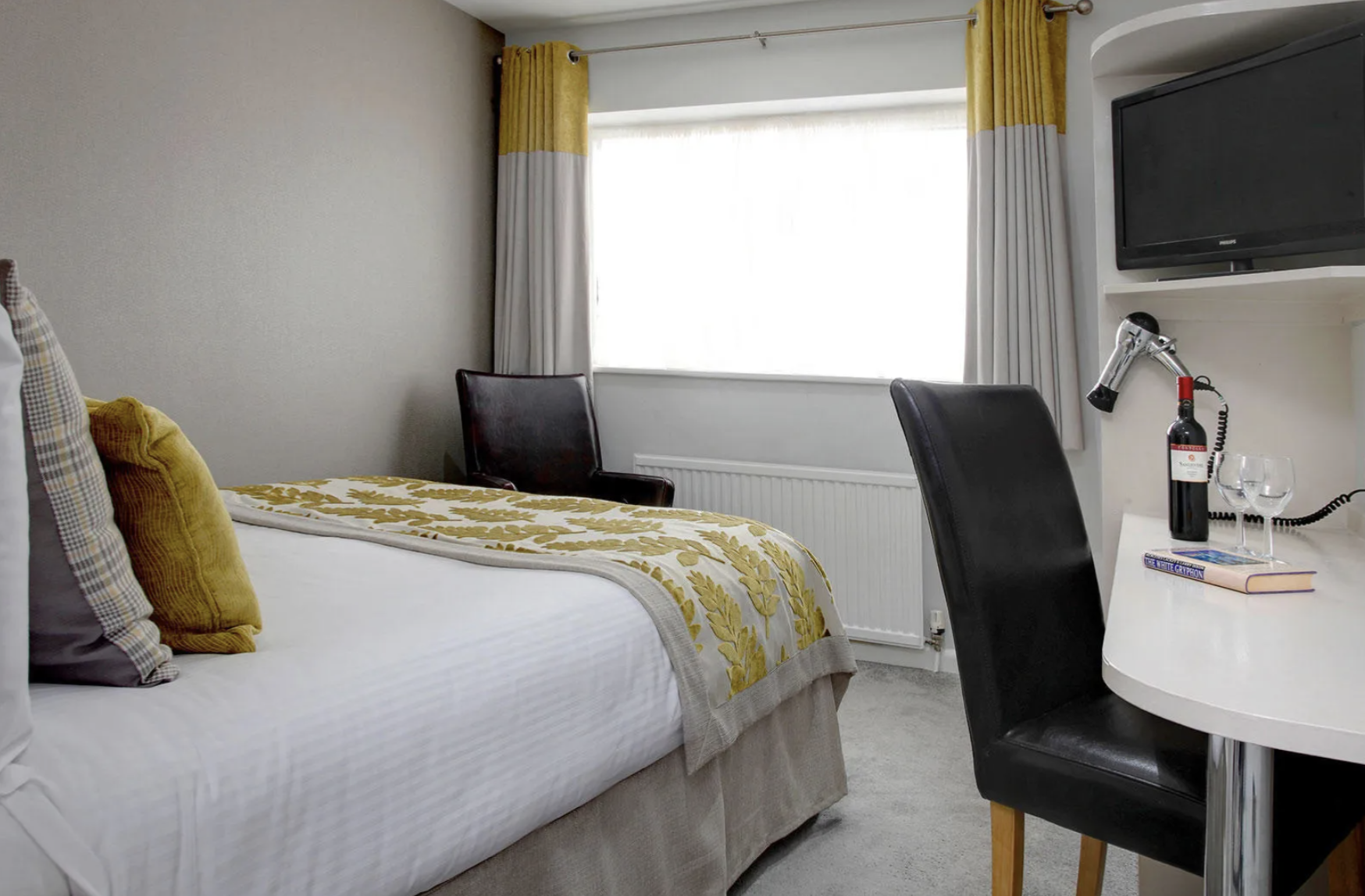 A hotel room with a bed, yellow curtains, a black chair by the window, a white desk with a bottle of wine, two wine glasses, a book, and a TV mounted on the wall.
