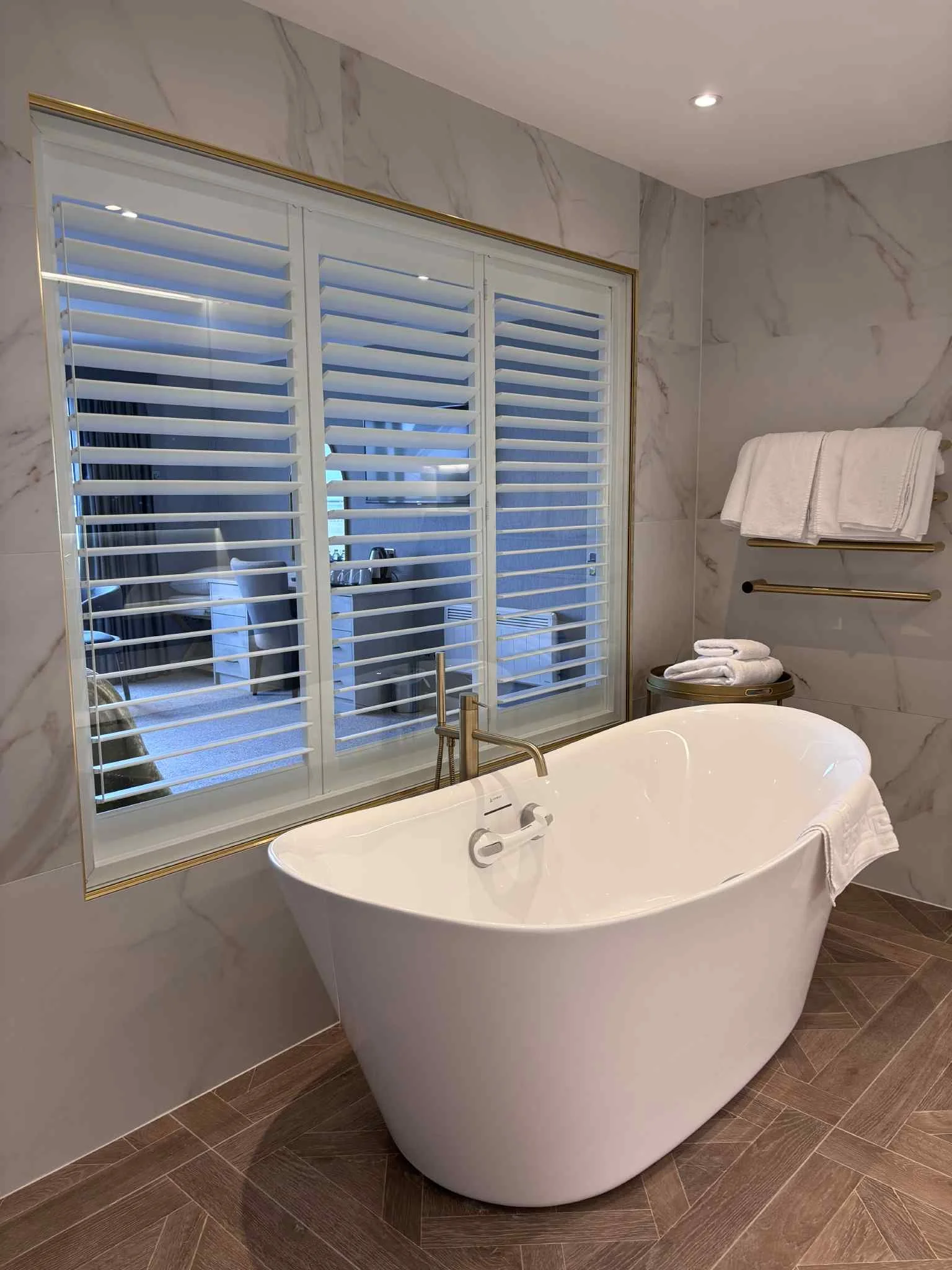 A luxurious bathroom with a freestanding white bathtub, a large window with white shutters, marble walls, wooden flooring, and towels on a wall-mounted shelf.