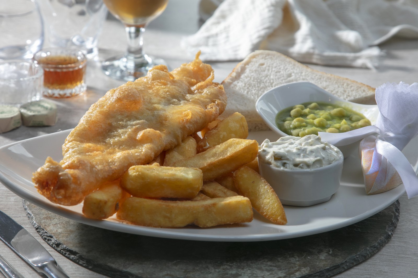 A plate of fish and chips with potato fries, a bowl of peas, and a container of tartar sauce on a table set for a meal.