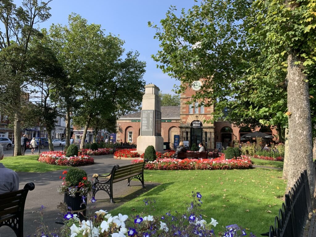 A park with flower beds, benches, and trees in front of a brick building with a clock and a monument in the center.