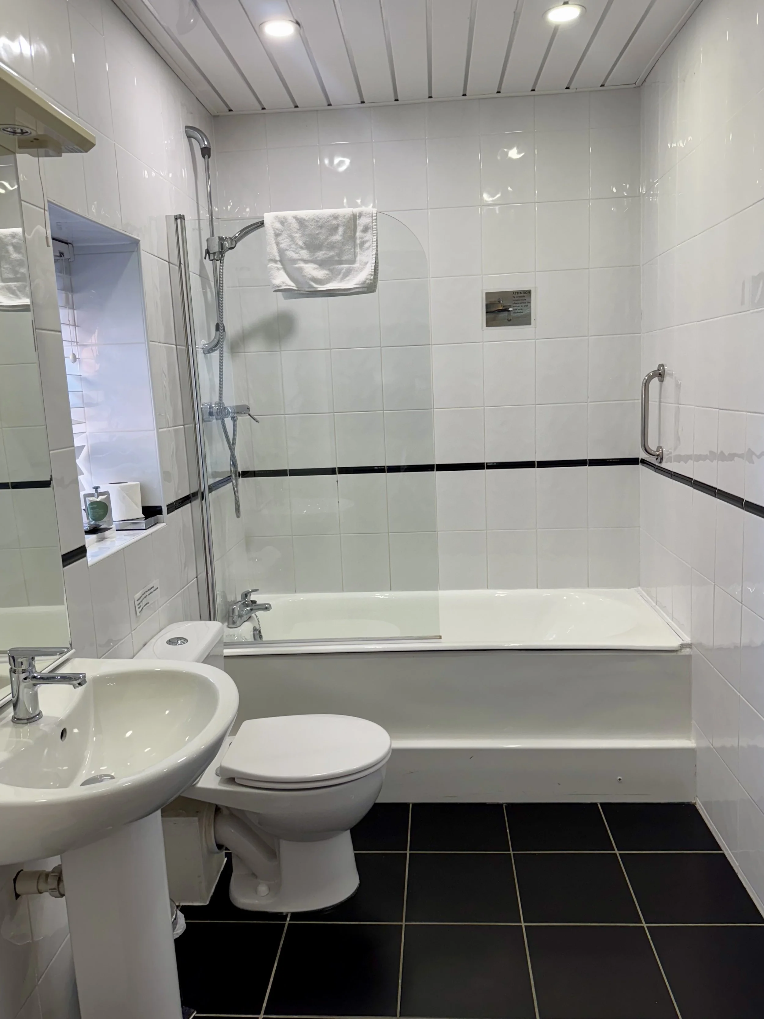 A small bathroom with white tiled walls, a bathtub with a glass divider, a toilet, and a pedestal sink. There is a shaving mirror with a shelf, a window with a small window sill, and a towel hanging on a shower rod. The floor is black with white grou