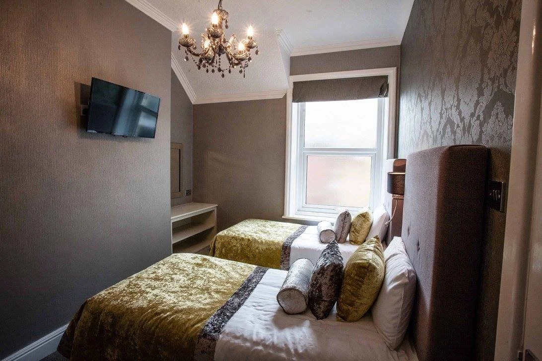 A bedroom with two single beds, each with green and gray bedspreads, multiple pillows, a window with gray shades, a TV mounted on a gray wall, and a chandelier hanging from the ceiling.