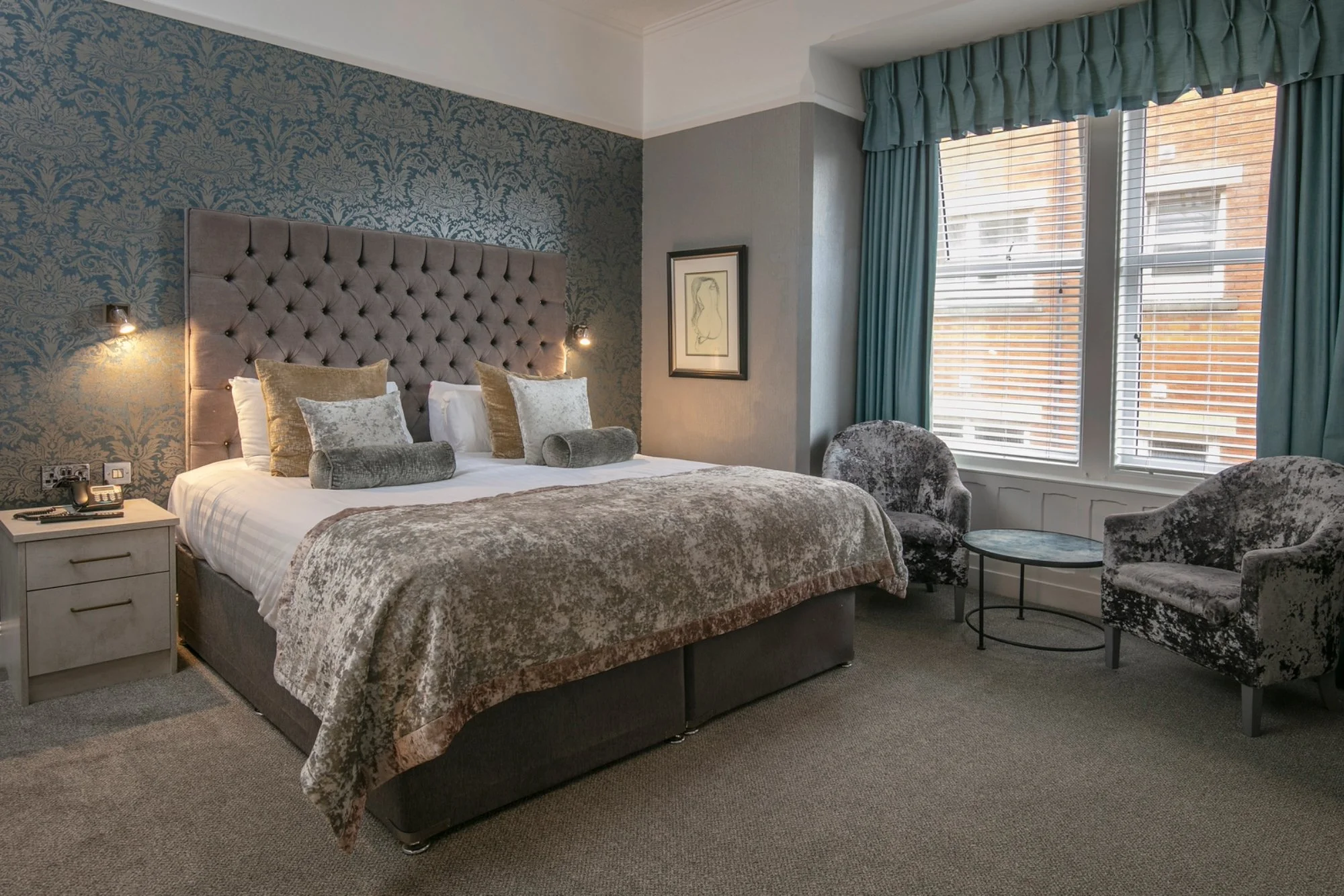 A hotel room with an upholstered bed, decorative pillows, bedside table with a phone, and window with blue curtains and white blinds.