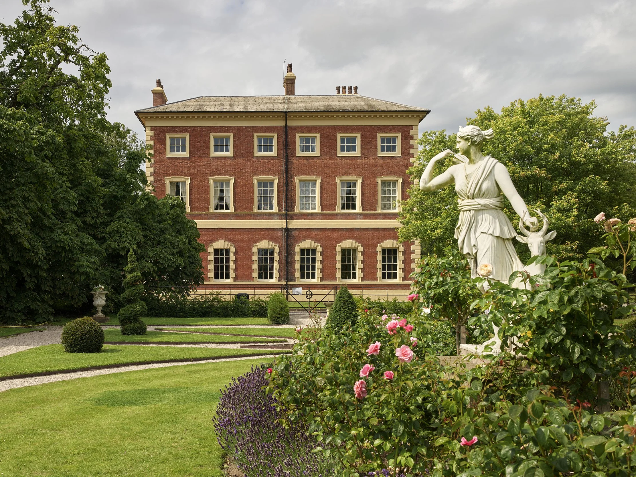 A garden with a classical statue of a woman and child, pink roses, green shrubs, and trees in front of a red brick historic building under a cloudy sky.