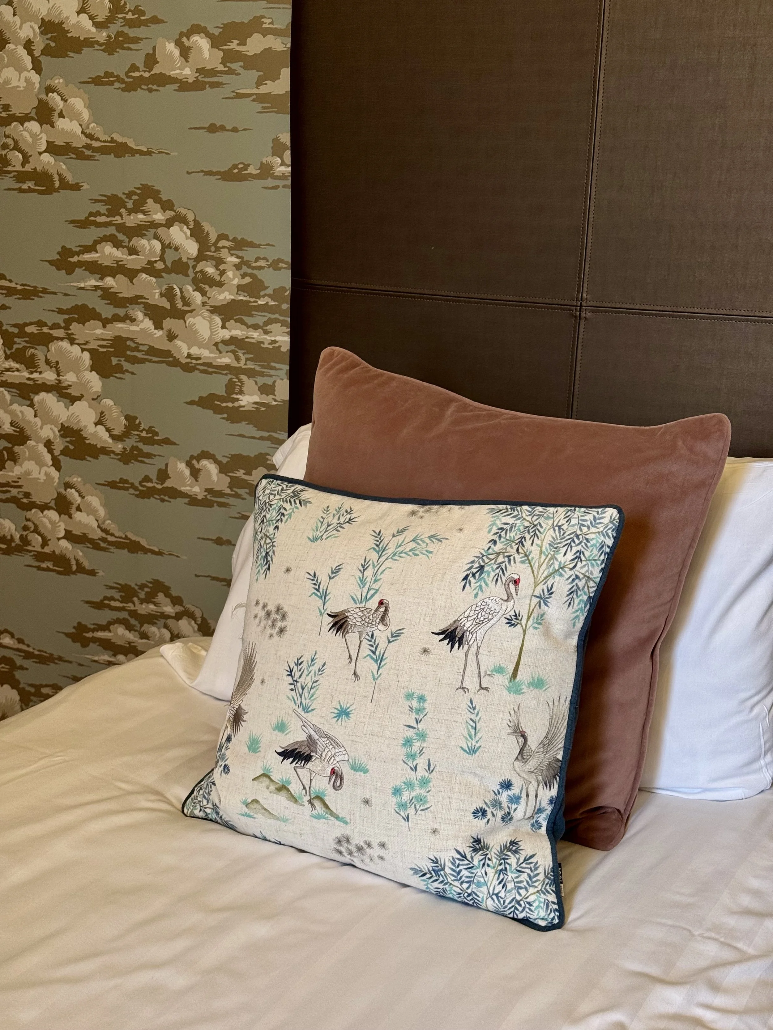 Decorative bed with embroidered pillow featuring cranes and foliage in front of a dark upholstered headboard and a wall with a cloud pattern.