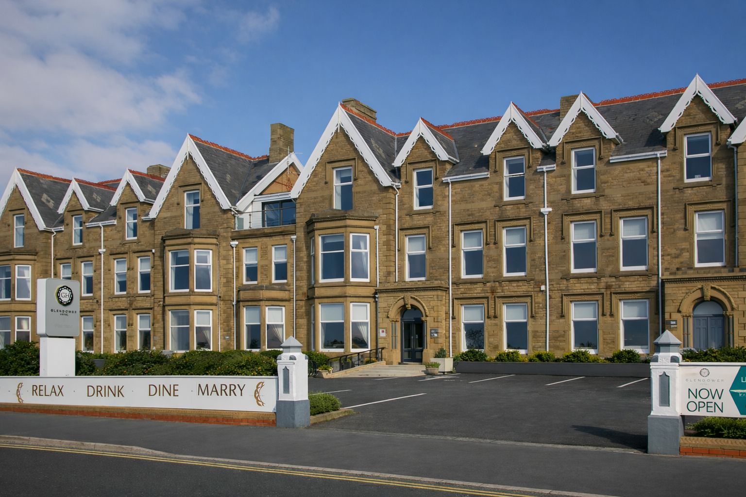 A historic stone building with bay windows, a sloped roof with decorative gable trim, and a parking lot in front. A sign indicates the Glendower Hotel property with a message to relax, drink, dine, and marry. The sky is partly cloudy but mostly blue.