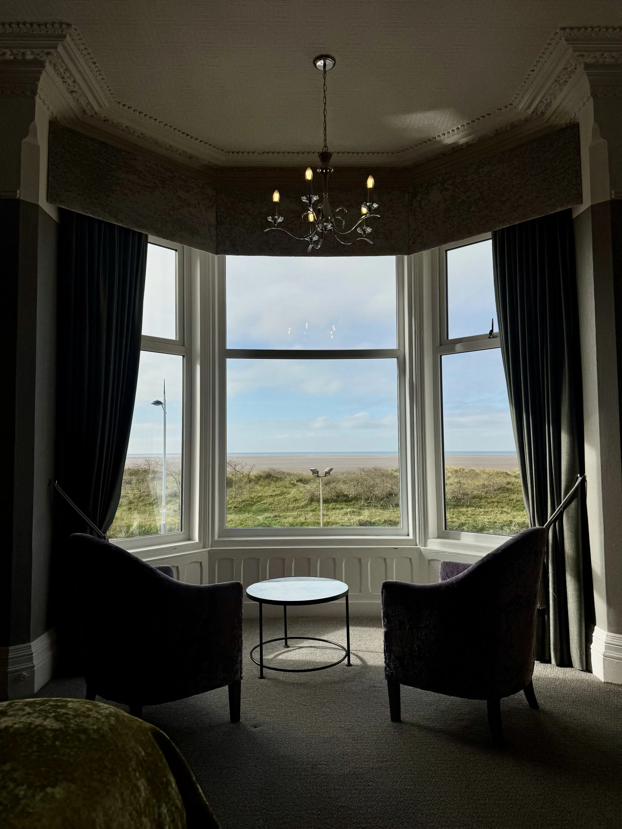 A cozy sitting area with two dark-colored armchairs facing a round table, set by a large bay window with a view of grassy dunes and the beach under a partly cloudy sky. Dark curtains frame the window, and a chandelier hangs from the ceiling.