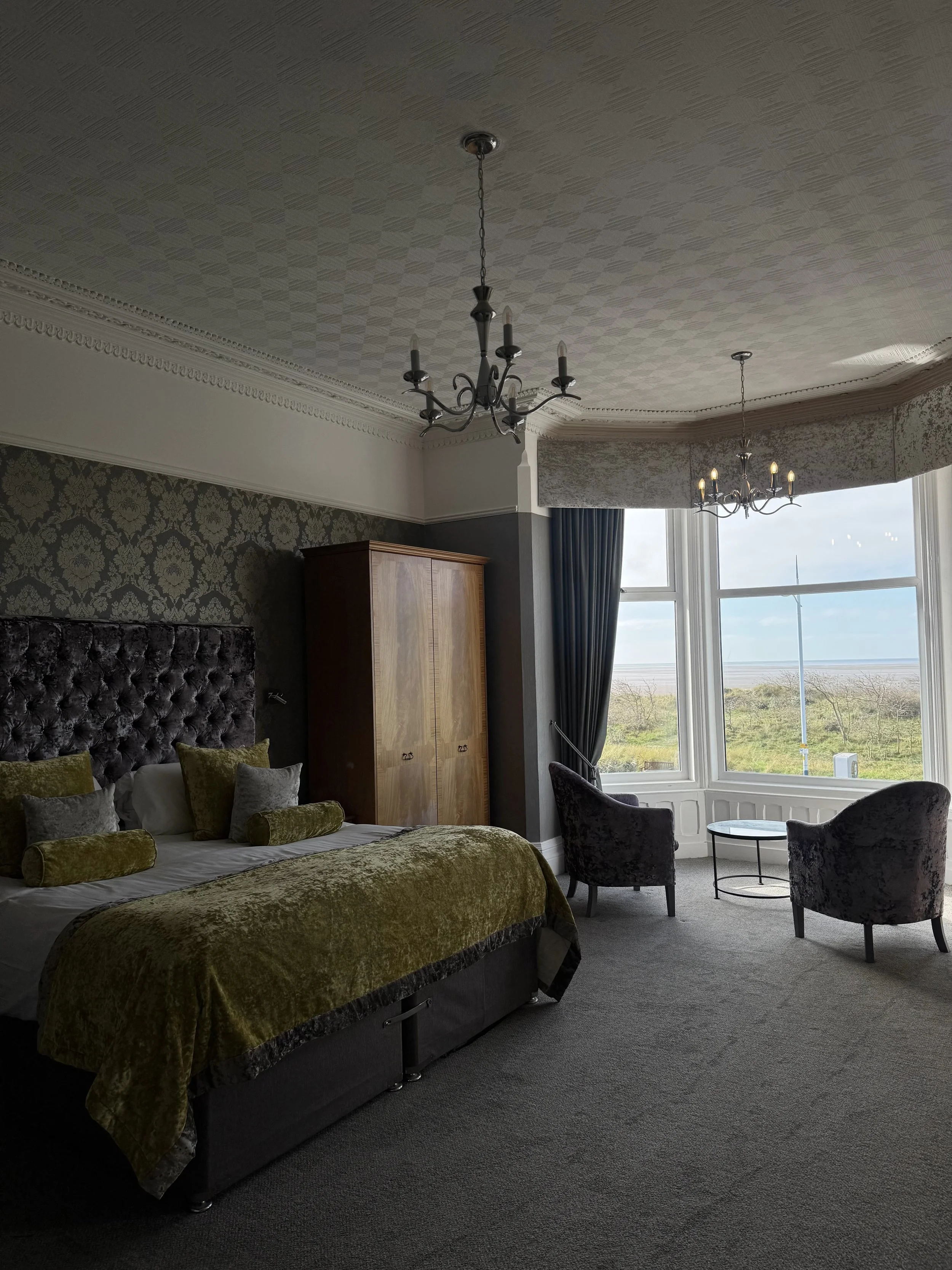 A bedroom with a large window featuring a view of grass and sky, two velvet armchairs, a side table, a tall cabinet, a bed with velvet bedding, and chandeliers hanging from the ceiling.