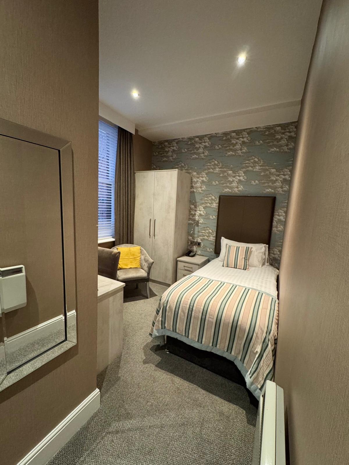 A cozy hotel room with a single bed, striped bedding, and a gray upholstered headboard, a white nightstand, a beige armchair with a yellow pillow, a white wardrobe, a window with brown curtains, and cloud-patterned wallpaper on one wall.