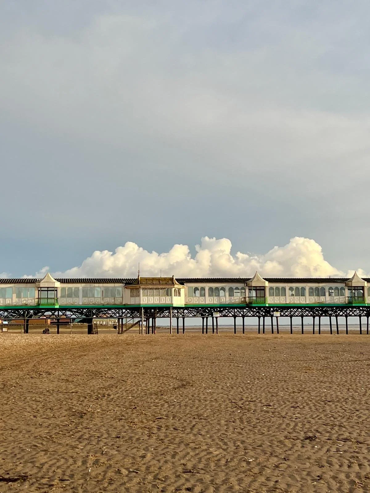 A pier extending over a sandy beach with a partly cloudy sky background.