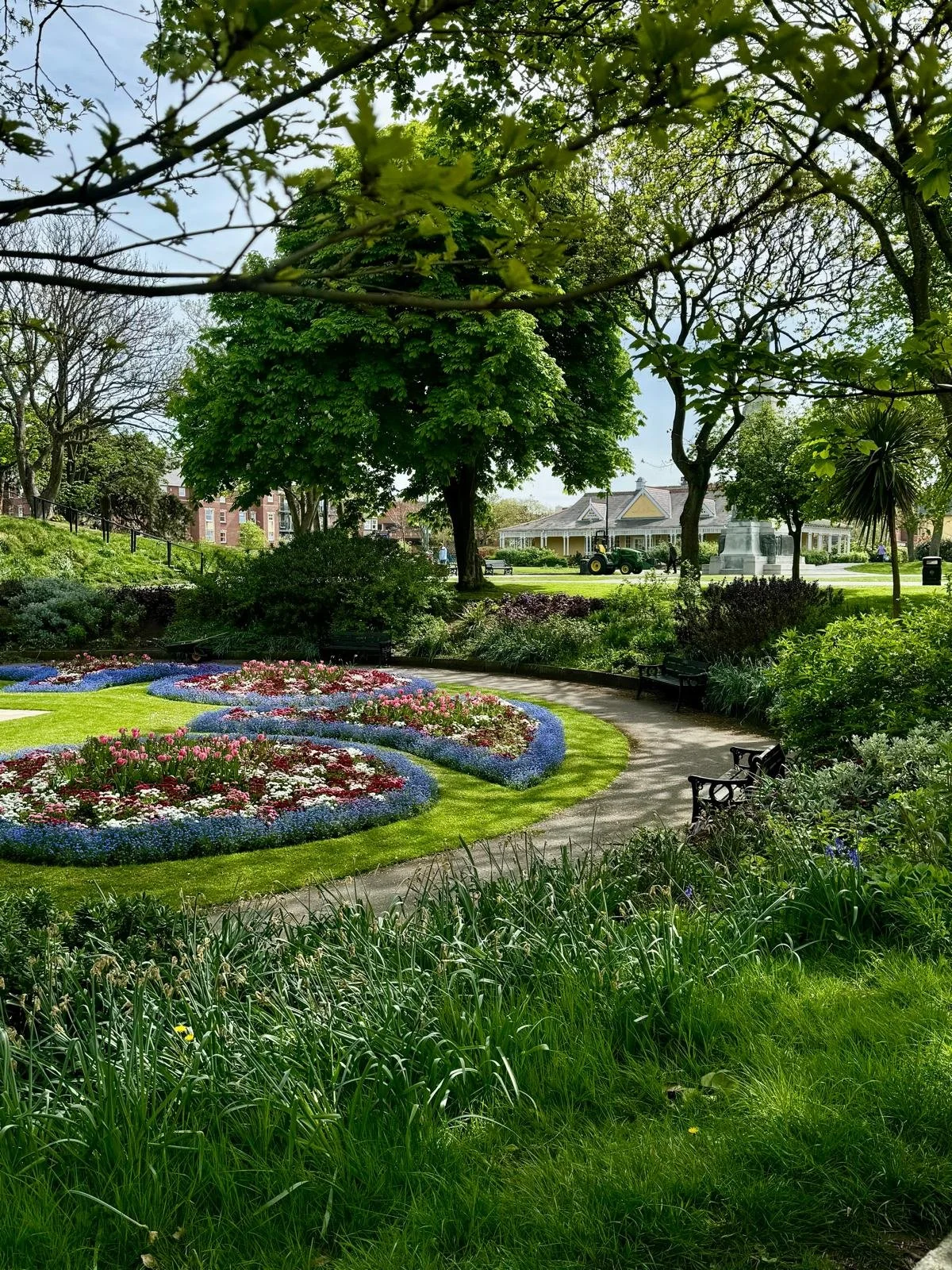 A lush park with flowering beds, a walking path, benches, and trees, with a gazebo and street scene in the background.