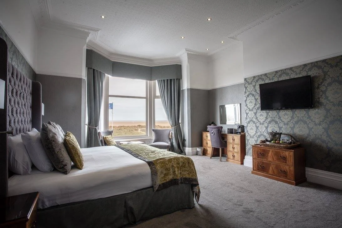 Luxury hotel room with a large bed, window with curtains showing the beach outside, a TV on the wall, and a vanity desk with a mirror.