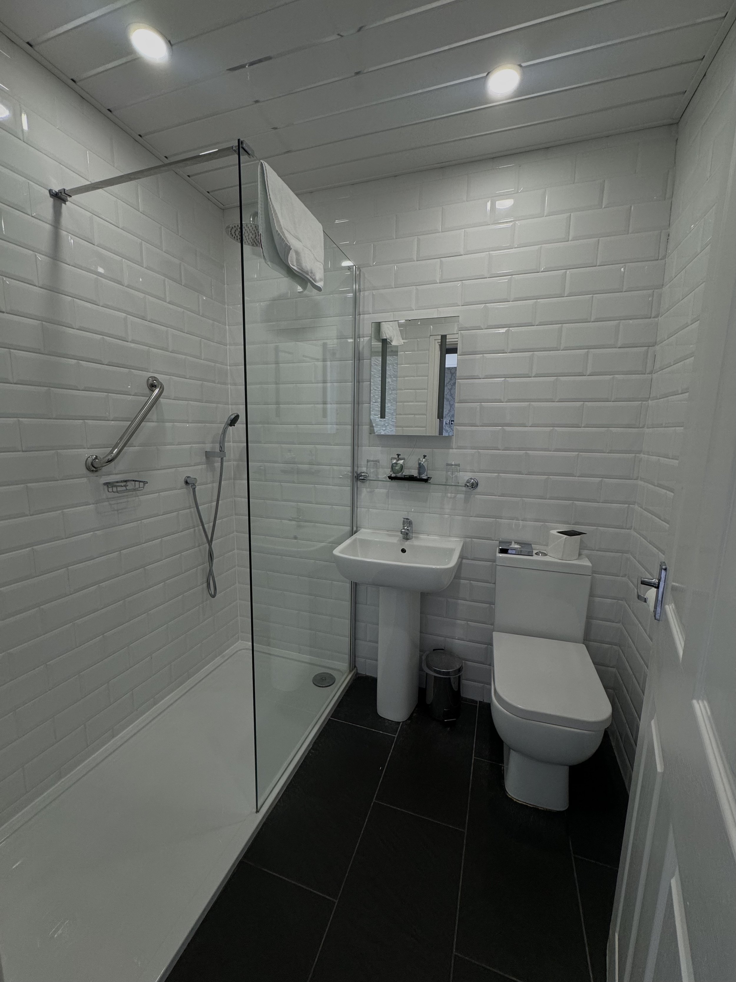 Small bathroom with white brick walls, a shower area with a glass partition, a pedestal sink, a mirror above the sink, a toilet, and a small trash bin. There are towel racks and toiletries on the sink and shelf.
