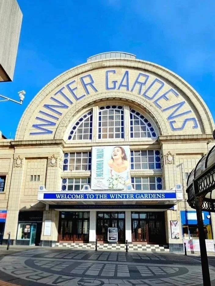 The entrance to Winter Gardens, a historic building with a large arched window and sign that reads 'Winter Gardens'. There is a banner with a woman and the text 'Winter Art'.
